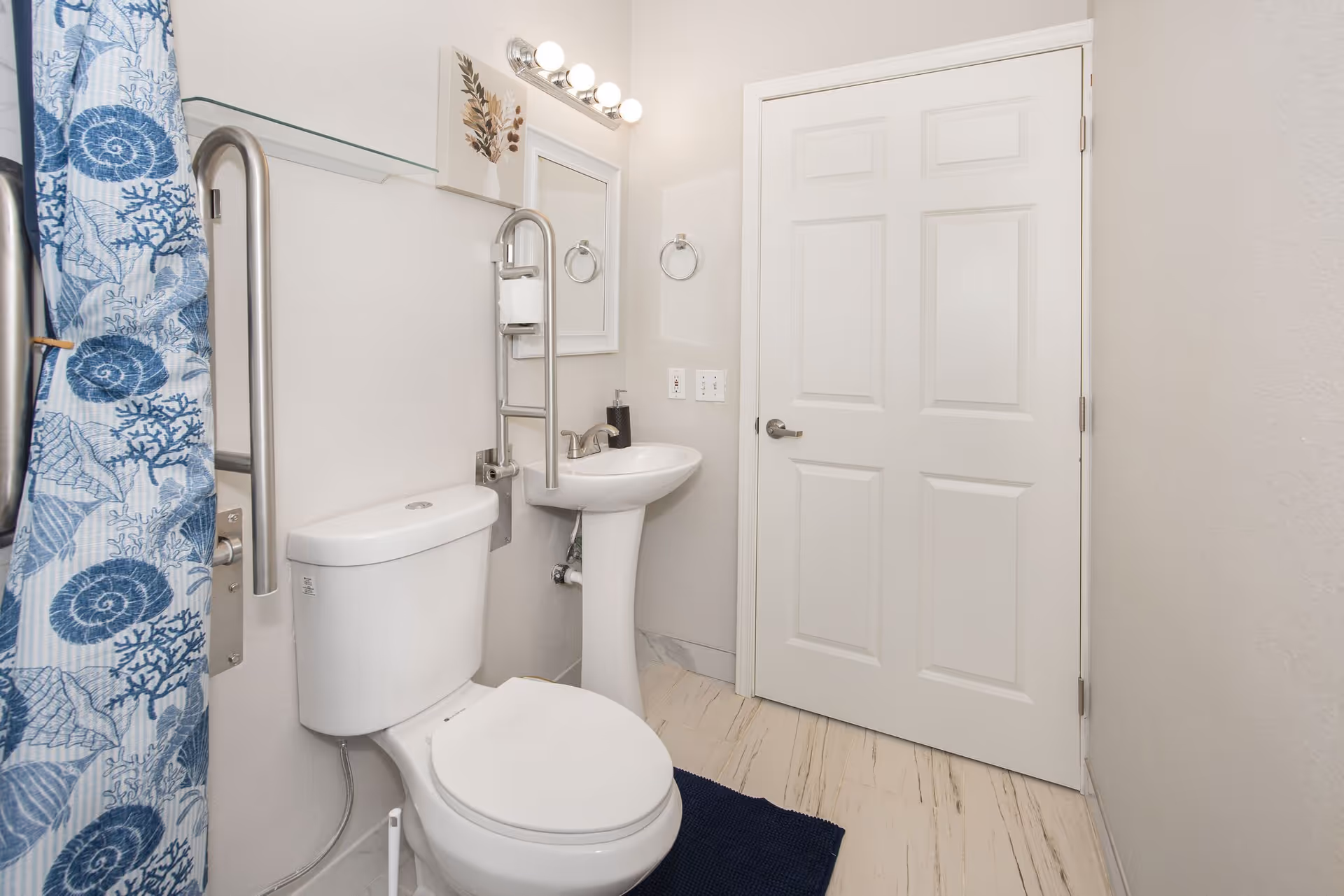 A clean bathroom with a white toilet, a white pedestal sink, a mirror with three light bulbs above it, a towel ring, and a blue and white shower curtain with seashell and coral patterns. The walls are light-colored, and the floor has light-colored tiles. There are stainless steel grab bars near the toilet and shower area.
