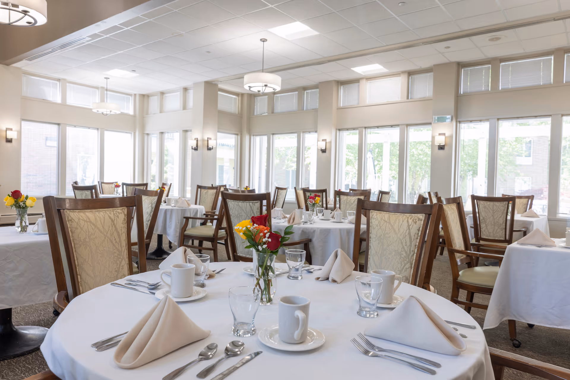 A bright and spacious dining room with multiple round tables covered in white tablecloths. Each table is set with folded beige napkins, white coffee cups, glasses, silverware, and small floral centerpieces. The room has large windows allowing natural light to fill the space, beige walls, and elegant wooden chairs with patterned upholstery.