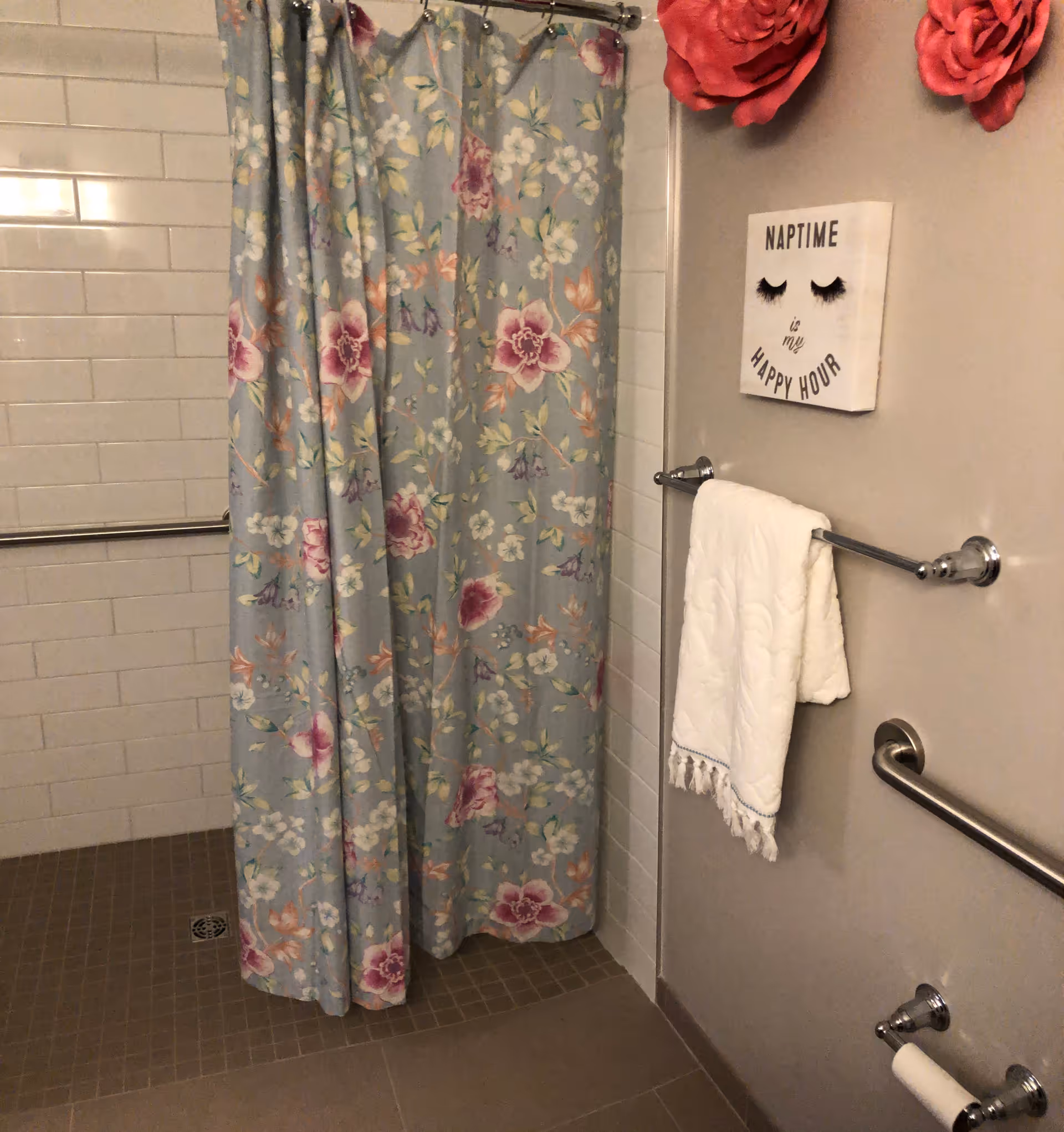 Bathroom with a floral shower curtain, white tiled walls, and brown tiled floor. A white towel hangs on a chrome towel rack attached to a beige wall. Above the towel rack is a decorative sign that reads 'NAPTIME is my HAPPY HOUR' with eyelash graphics. There are also two large red flower decorations on the wall.