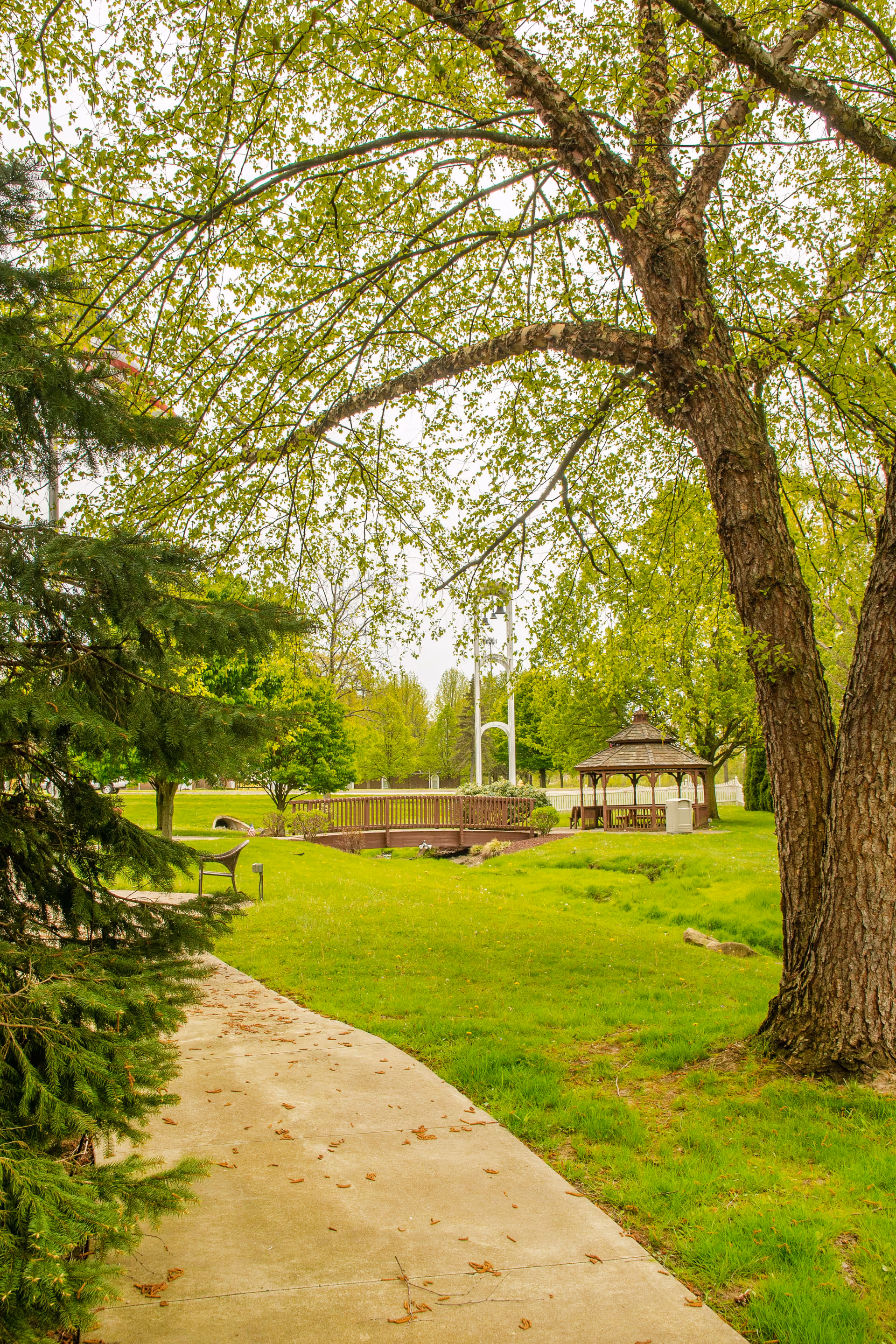 A peaceful outdoor garden area with a concrete pathway leading towards a wooden gazebo and a small wooden bridge. The scene is surrounded by lush green grass, trees with fresh green leaves, and a bench along the path.