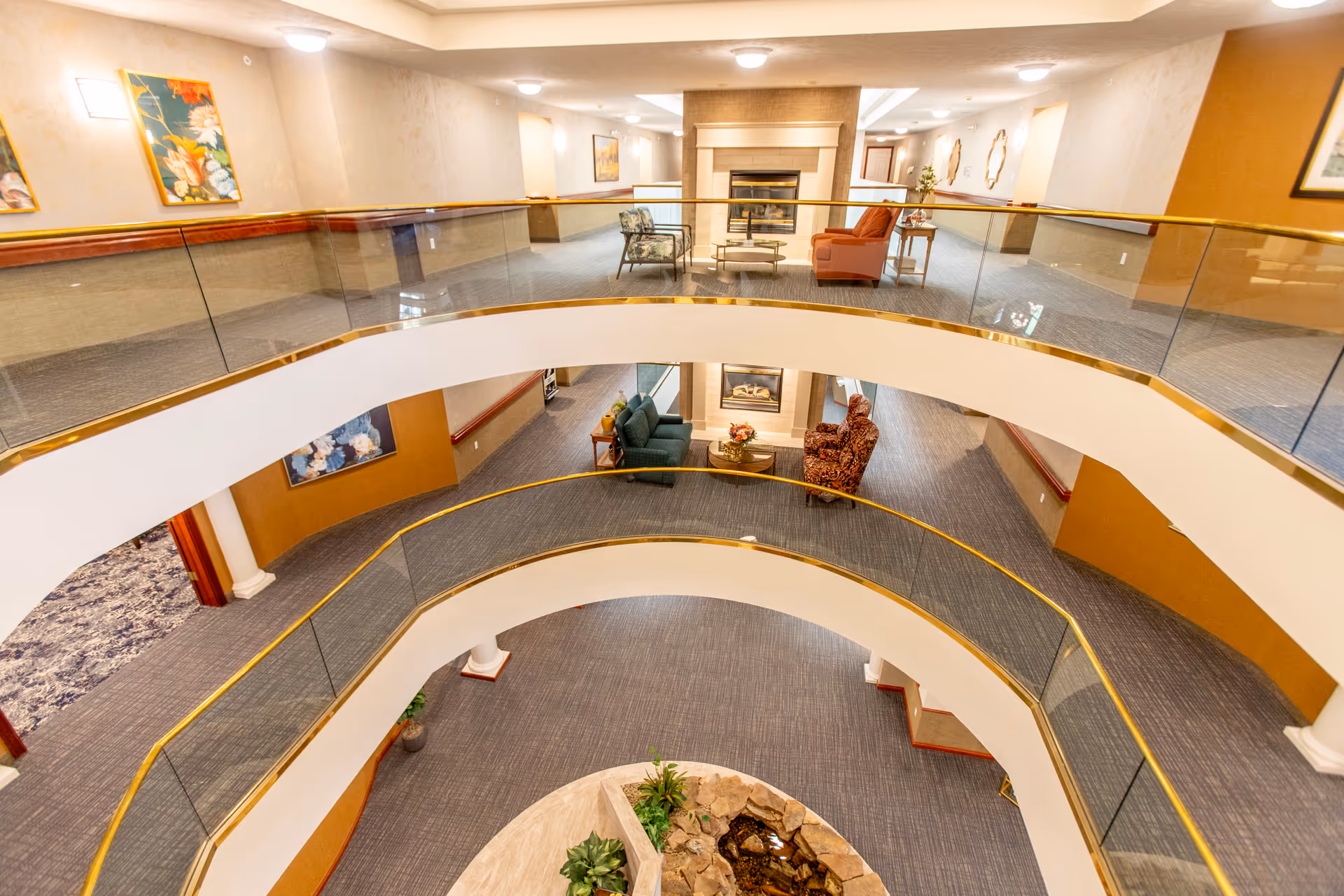 Three-level atrium inside a retirement community with curved glass-and-brass railings, seating areas, a fireplace, and indoor plants.