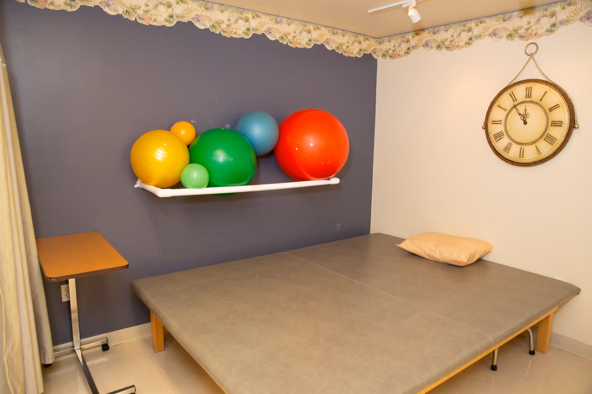 A therapy room with a padded treatment table and pillow, colorful exercise balls on a wall shelf, a small rolling table, and a clock on the wall.