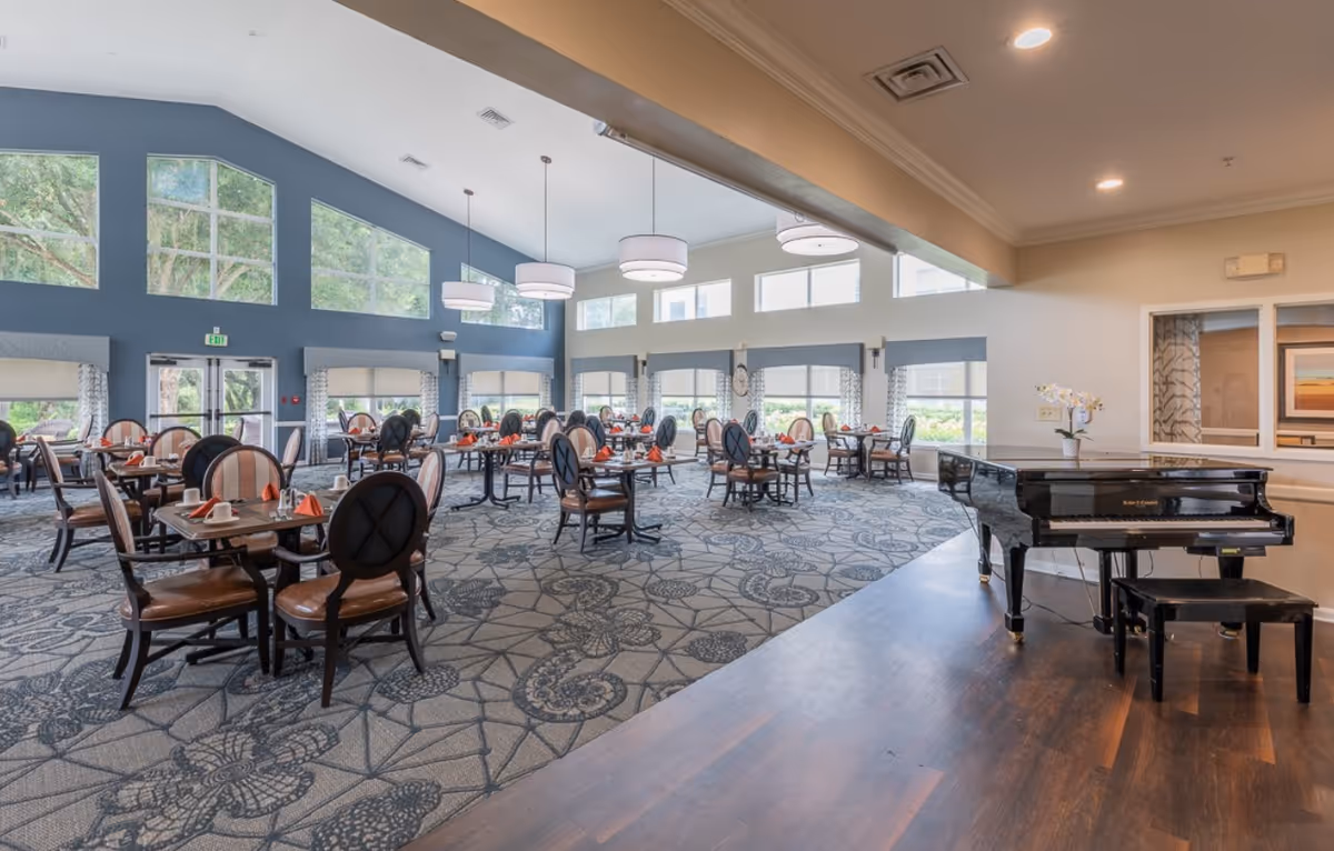 Spacious dining room with multiple round tables and chairs, large windows and pendant lights, and a black grand piano on a raised wooden platform.