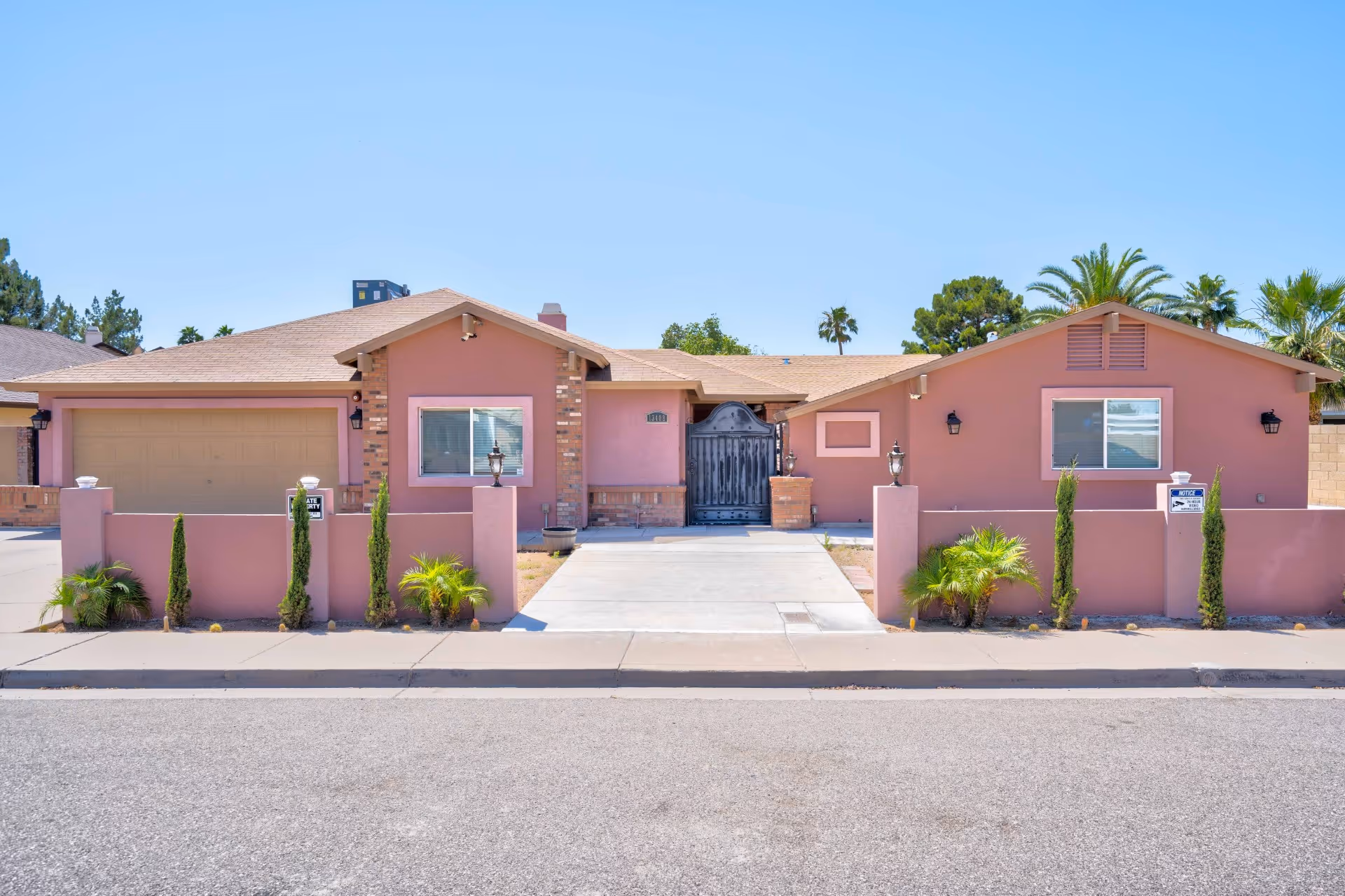 Front exterior view of a single-story pink care home building with a driveway, a garage, a gated entrance, small plants, and a clear blue sky.