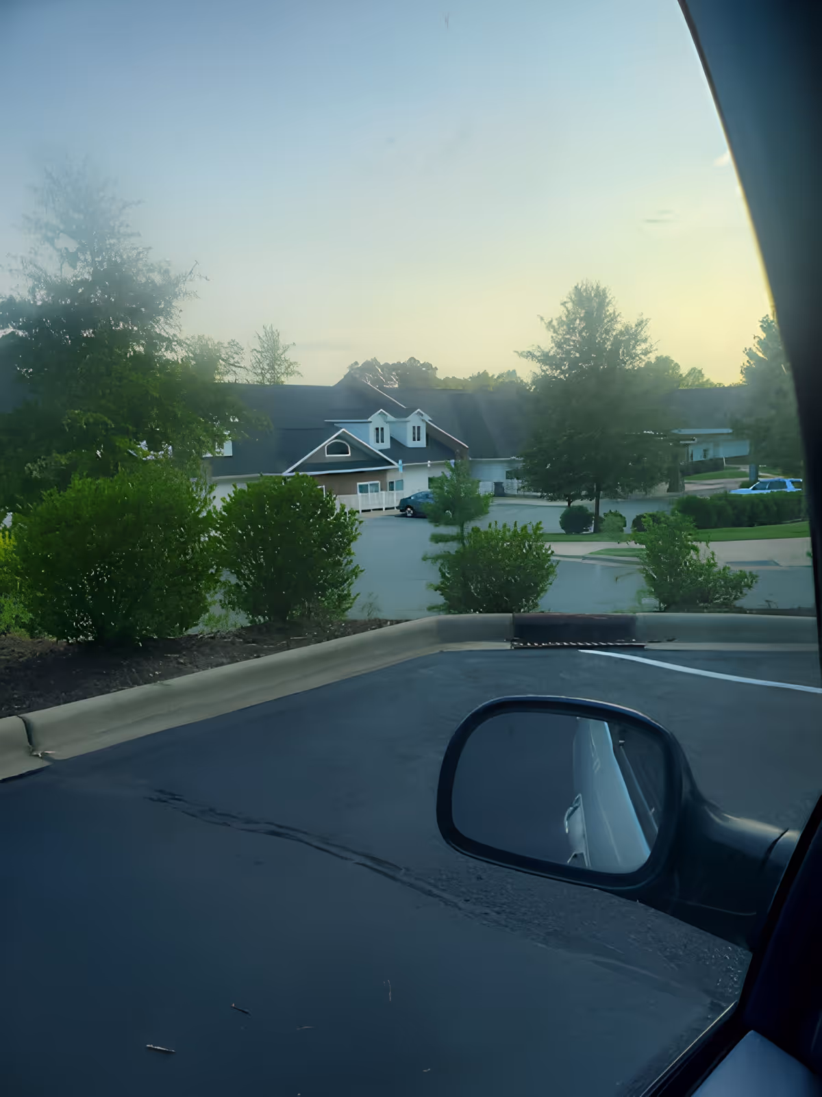 View from inside a parked car showing the side mirror, a parking lot and curb, landscaping, and a multi-gabled building with trees beyond.