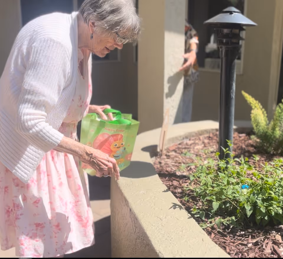 An elderly woman wearing a white cardigan and a pink floral dress is outdoors, holding a green Easter-themed bag and leaning over a low wall near a garden bed with plants and a black outdoor lamp post.