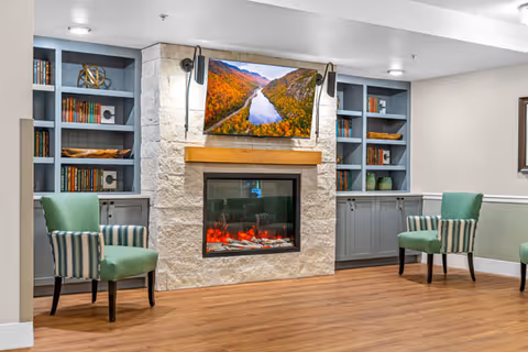 A cozy living room area featuring a stone fireplace with an electric fire, a flat-screen TV mounted above displaying a scenic river and autumn trees. On either side of the fireplace are built-in blue-gray bookshelves with books and decorative items. Two green upholstered chairs with striped armrests are positioned facing the fireplace on a wooden floor.