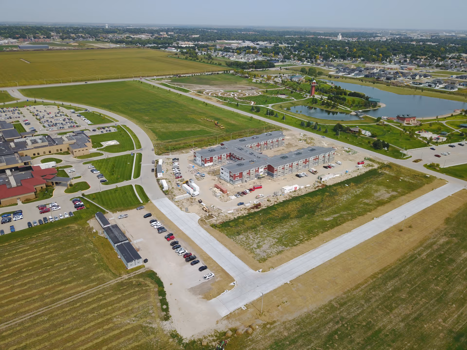 Aerial view of a large senior living facility complex under construction surrounded by green fields, parking lots, roads, and a landscaped area with a pond and a lighthouse structure in the background.