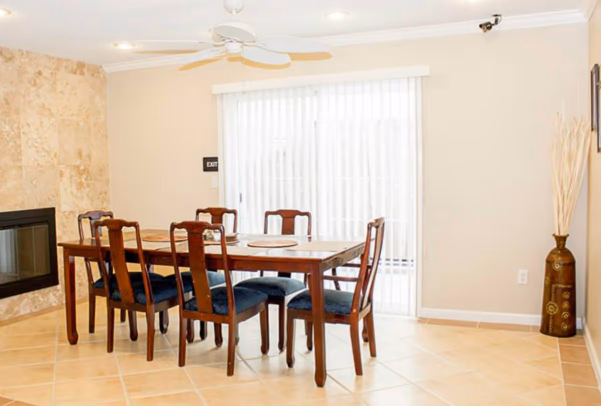 A dining area with a wooden table and six chairs with dark blue cushions. The room has beige tiled flooring, a ceiling fan, a stone fireplace on the left wall, and vertical blinds covering a sliding glass door in the background. There is a decorative vase with tall dried plants in the corner on the right.