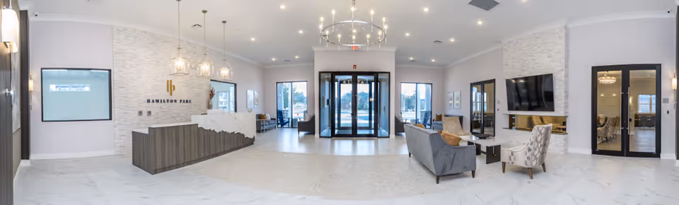 Spacious and modern senior living facility lobby with a reception desk on the left featuring the Hamilton Parc logo on a white textured wall. The lobby has large glass entrance doors in the center, seating areas with sofas and armchairs on the right, a mounted flat-screen TV above a fireplace, and large windows allowing natural light to fill the space. The floor is a mix of white marble and light-colored tiles, and the ceiling has elegant chandeliers and recessed lighting.