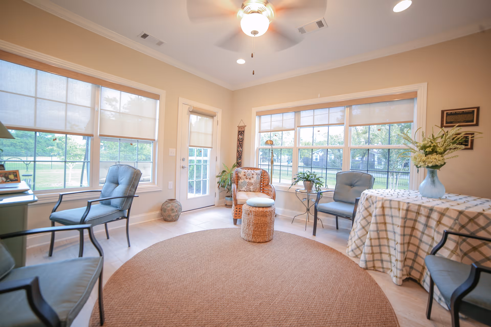 A bright and cozy sitting room with large windows letting in natural light. The room features a round woven rug, several cushioned chairs, a wicker chair with a small round ottoman, a table covered with a checkered cloth holding a vase of flowers, and some potted plants near the windows. A ceiling fan with a light is mounted on the ceiling.