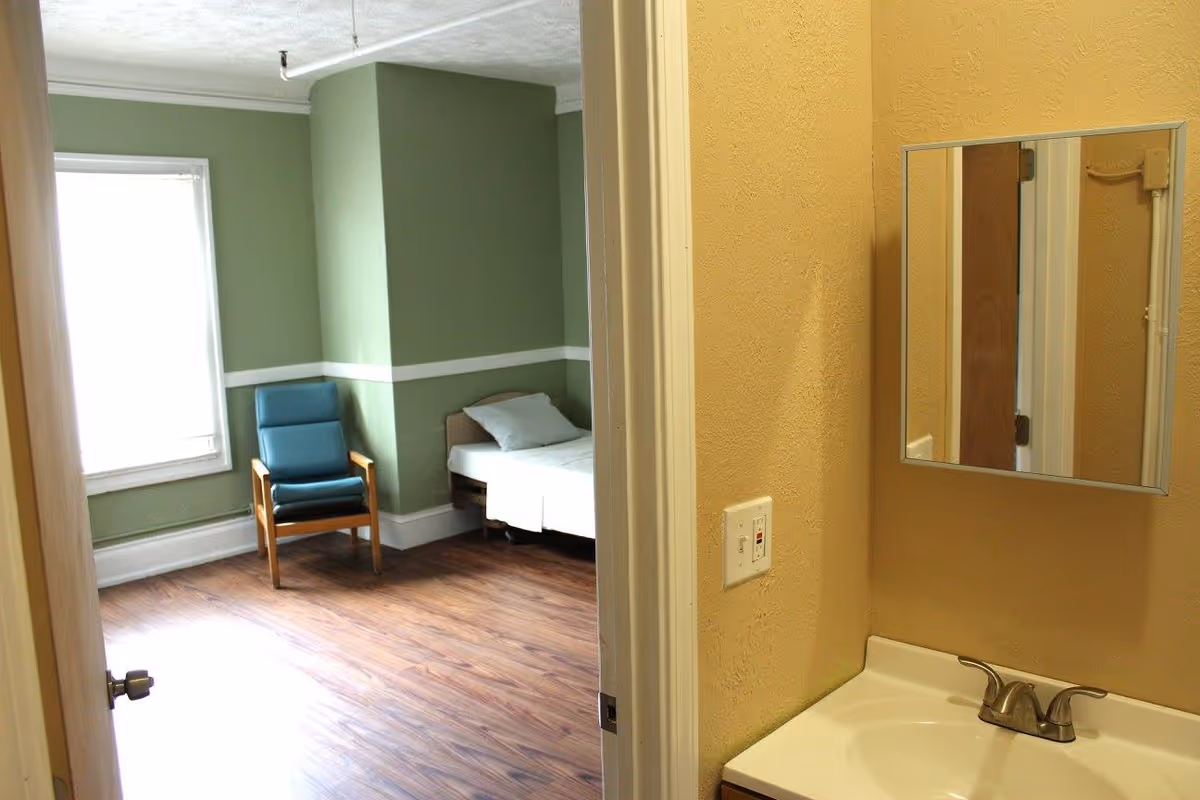 View into a small bedroom with green walls, a single bed and a blue armchair, seen from a beige washroom area with sink and mirror.