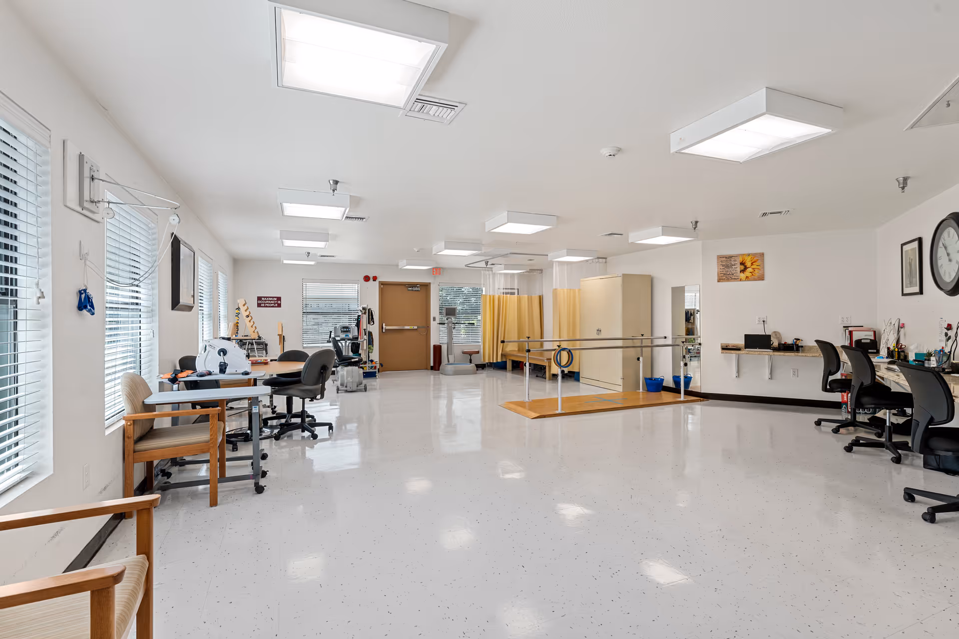 A spacious rehabilitation and healthcare room with white walls and ceiling, bright overhead lights, and large windows with blinds. The room contains various chairs, desks, exercise equipment, parallel bars for physical therapy, and a few privacy curtains. There are clocks and framed pictures on the walls, and the floor is a clean, shiny white surface.