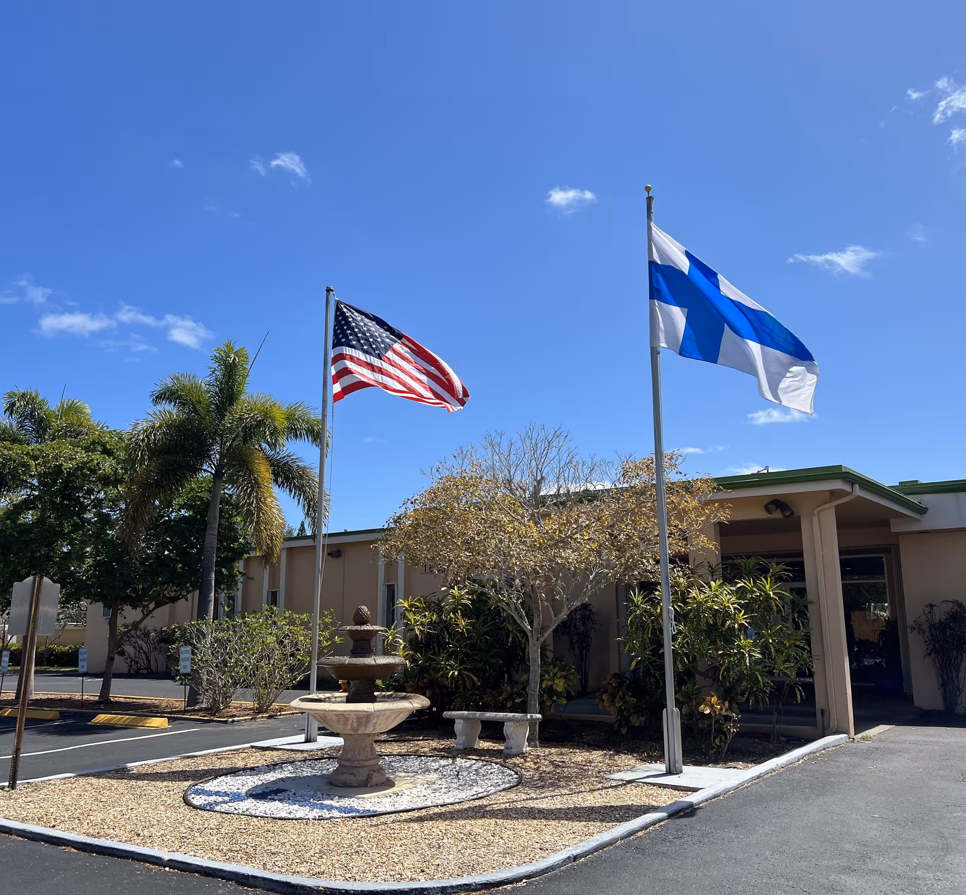 Exterior view of Finnish-American Village facility with two flagpoles displaying the American flag and the Finnish flag. There is a small fountain surrounded by gravel and a bench in front of the building. Palm trees and other greenery are visible under a clear blue sky.