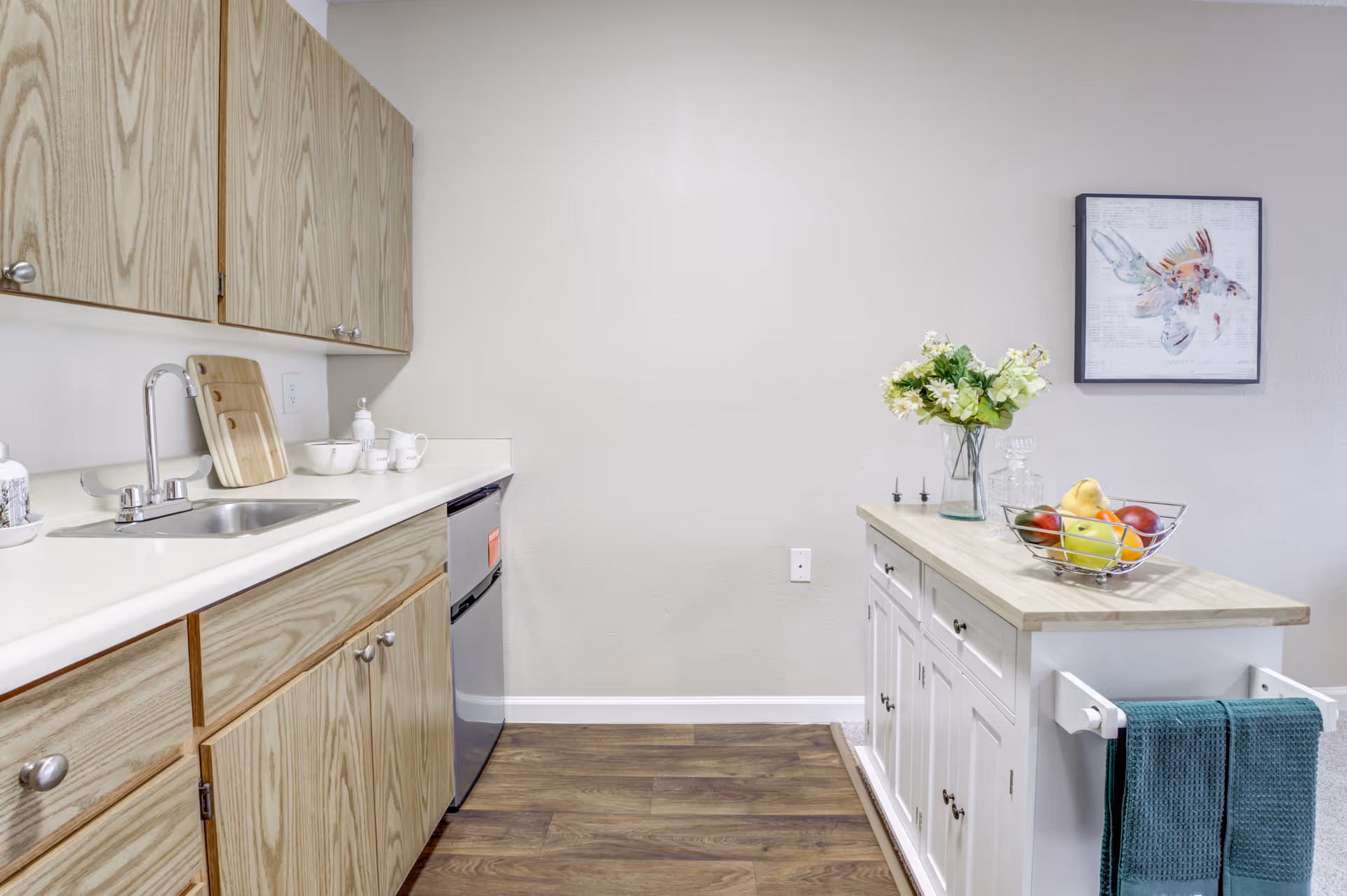 A clean and modern kitchen area with wooden cabinets, a stainless steel sink, and a small dishwasher. There is a kitchen island with a wooden countertop, decorated with a vase of flowers and a bowl of fruit. A framed picture of a fish hangs on the wall above the island. The floor is wooden and there are two teal towels hanging on the side of the island.