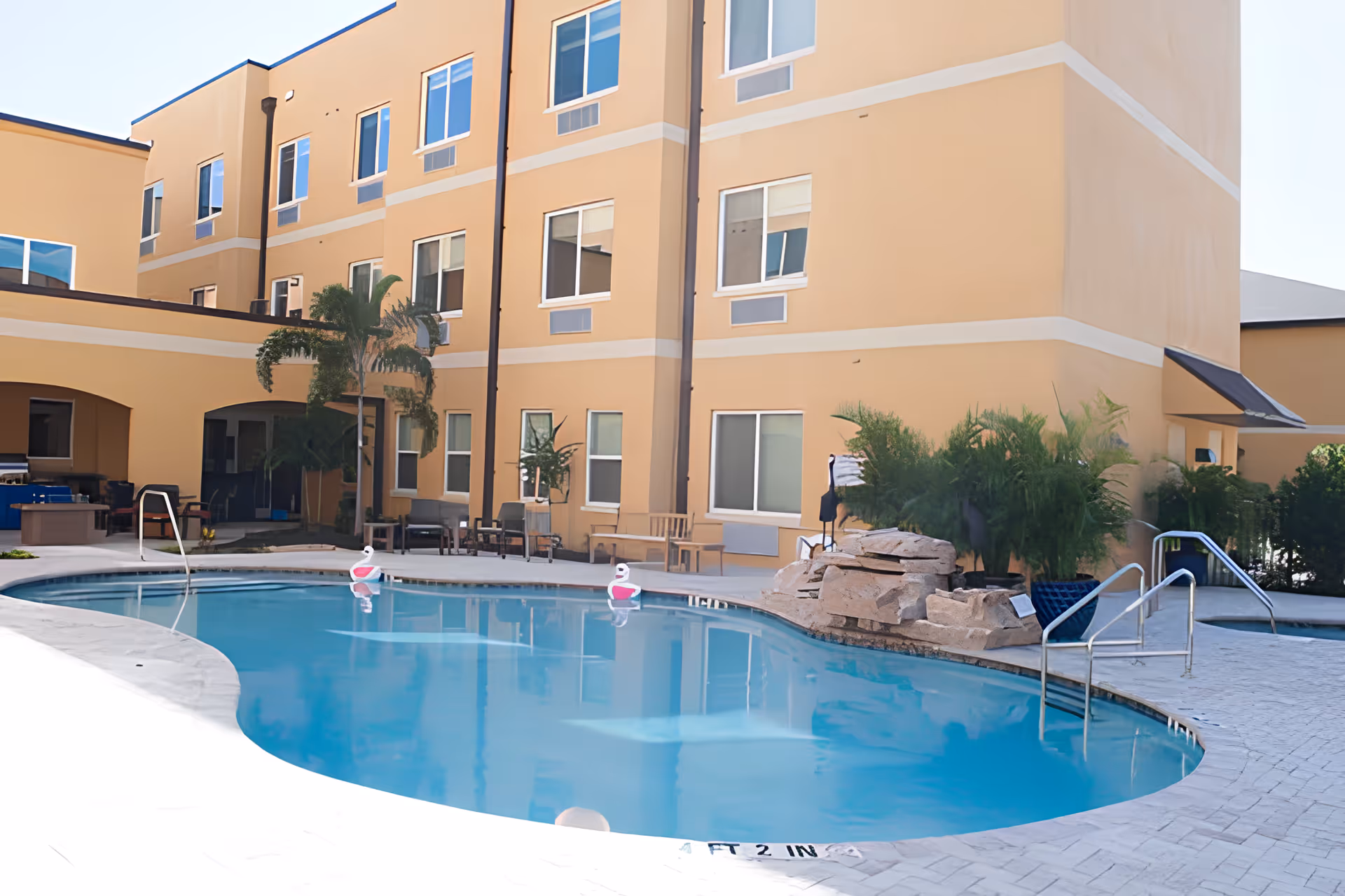 Outdoor swimming pool area at Aravilla Sarasota with clear blue water, surrounded by a beige building with multiple windows. There are poolside chairs, tables, palm trees, and some greenery around the pool. The pool has metal handrails and a rock feature near one side.