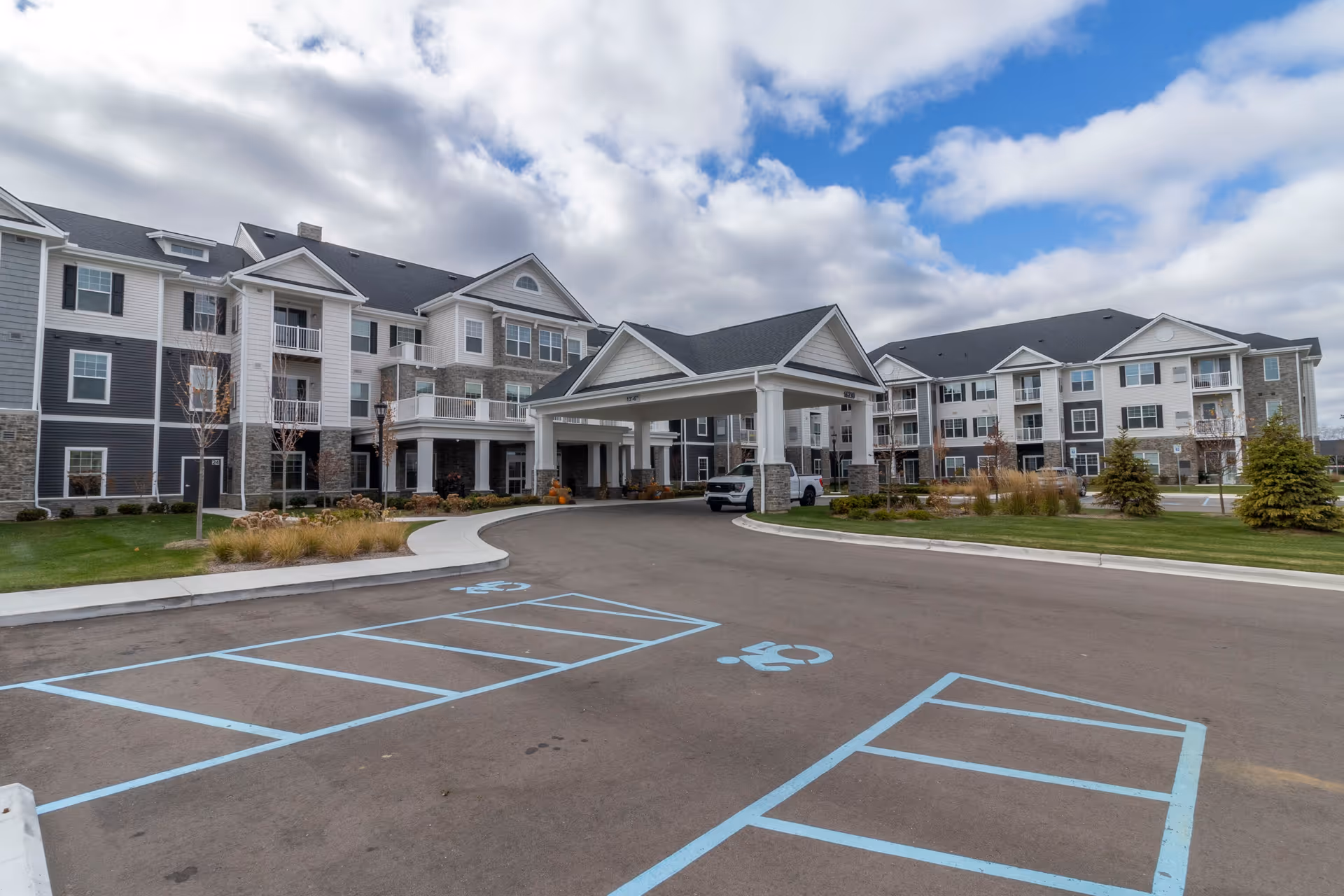 Exterior view of a senior living facility with a covered entrance driveway, multiple three-story buildings with balconies and windows, landscaped greenery, and a partly cloudy sky.