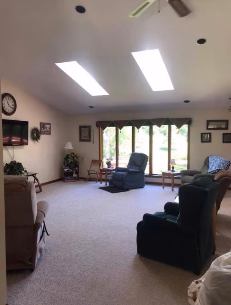 A spacious living room with beige carpet and multiple recliner chairs arranged facing a large window with a view of greenery outside. The room has two rectangular skylights on the ceiling, a wall-mounted TV, a clock, framed pictures, and small tables with lamps and plants.