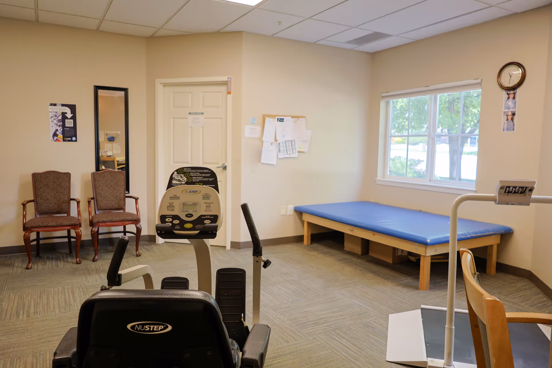 A small interior therapy room with exercise equipment (NuStep), a blue treatment table, two chairs, bulletin board and a window.