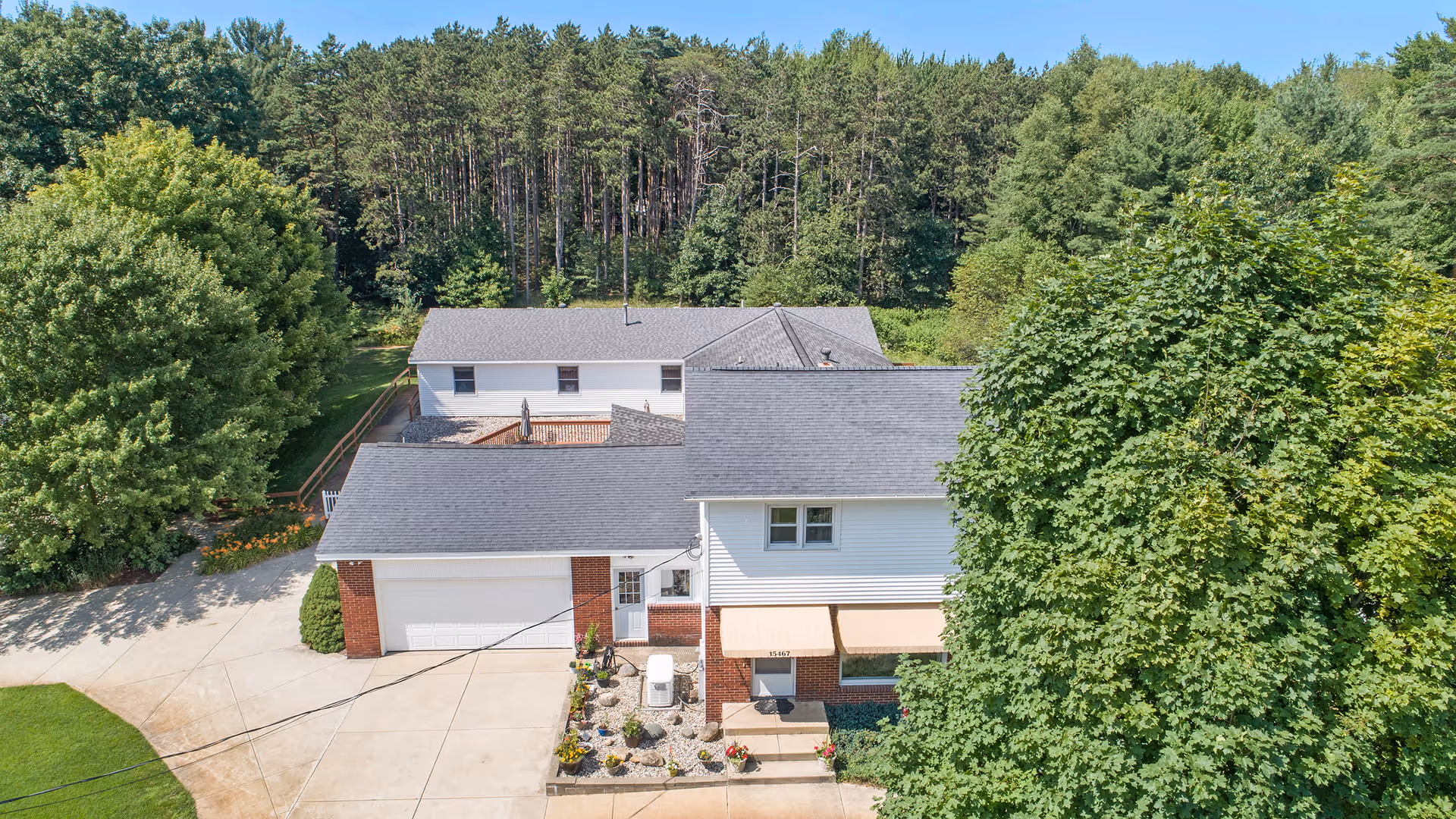 Aerial view of Pine Ridge Assisted Living facility showing a white and brick building with a gray roof surrounded by large green trees and a driveway. The building is set against a backdrop of dense forest under a clear blue sky.
