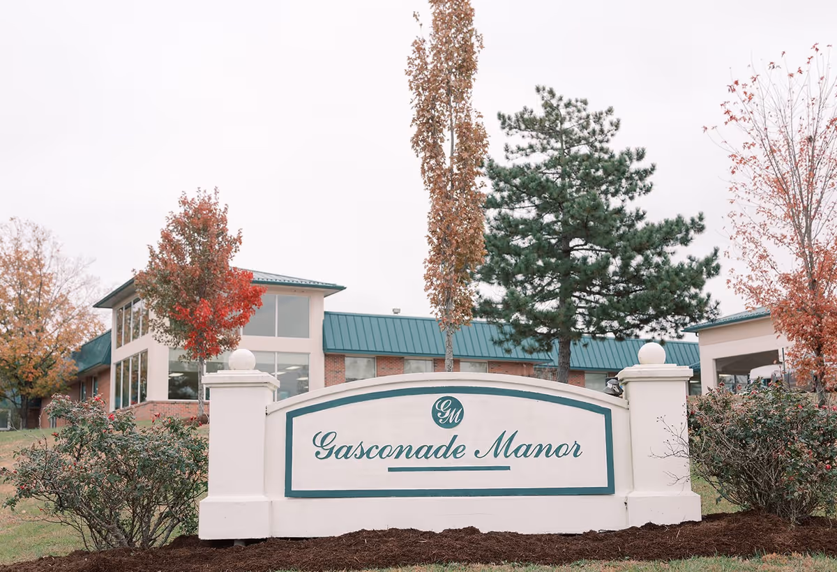 Entrance sign reading "Gasconade Manor" in front of a brick nursing home building with trees and a green roof.