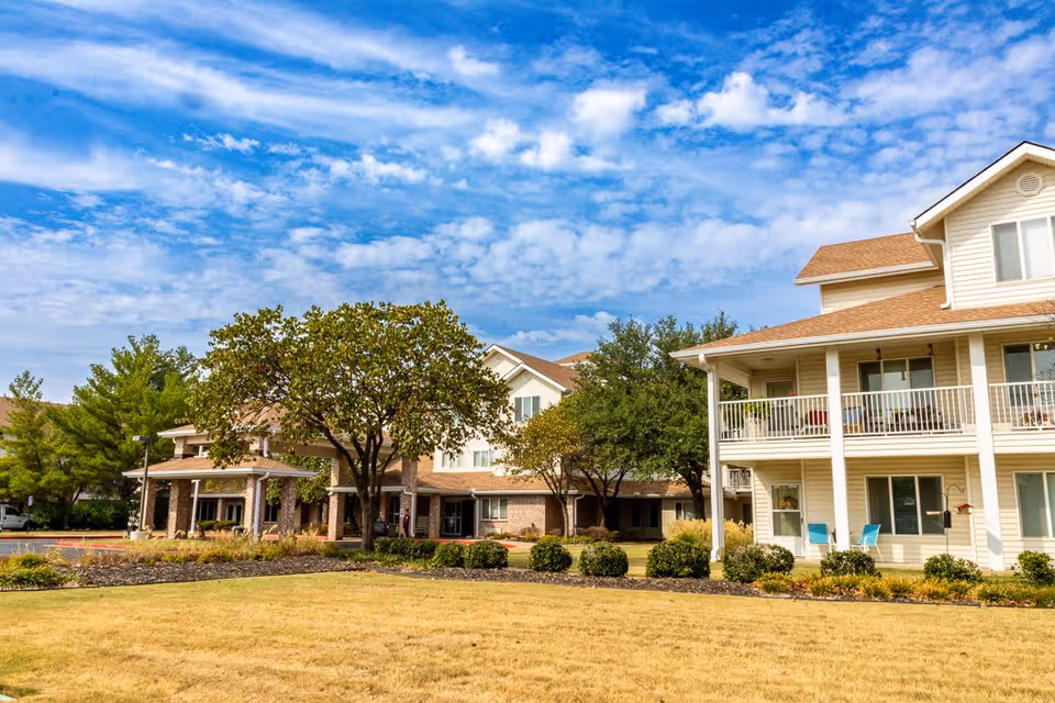 Exterior view of Asher Point Independent Living of Bartlesville showing a multi-story residential building with balconies, surrounded by trees and landscaped grounds under a partly cloudy blue sky.
