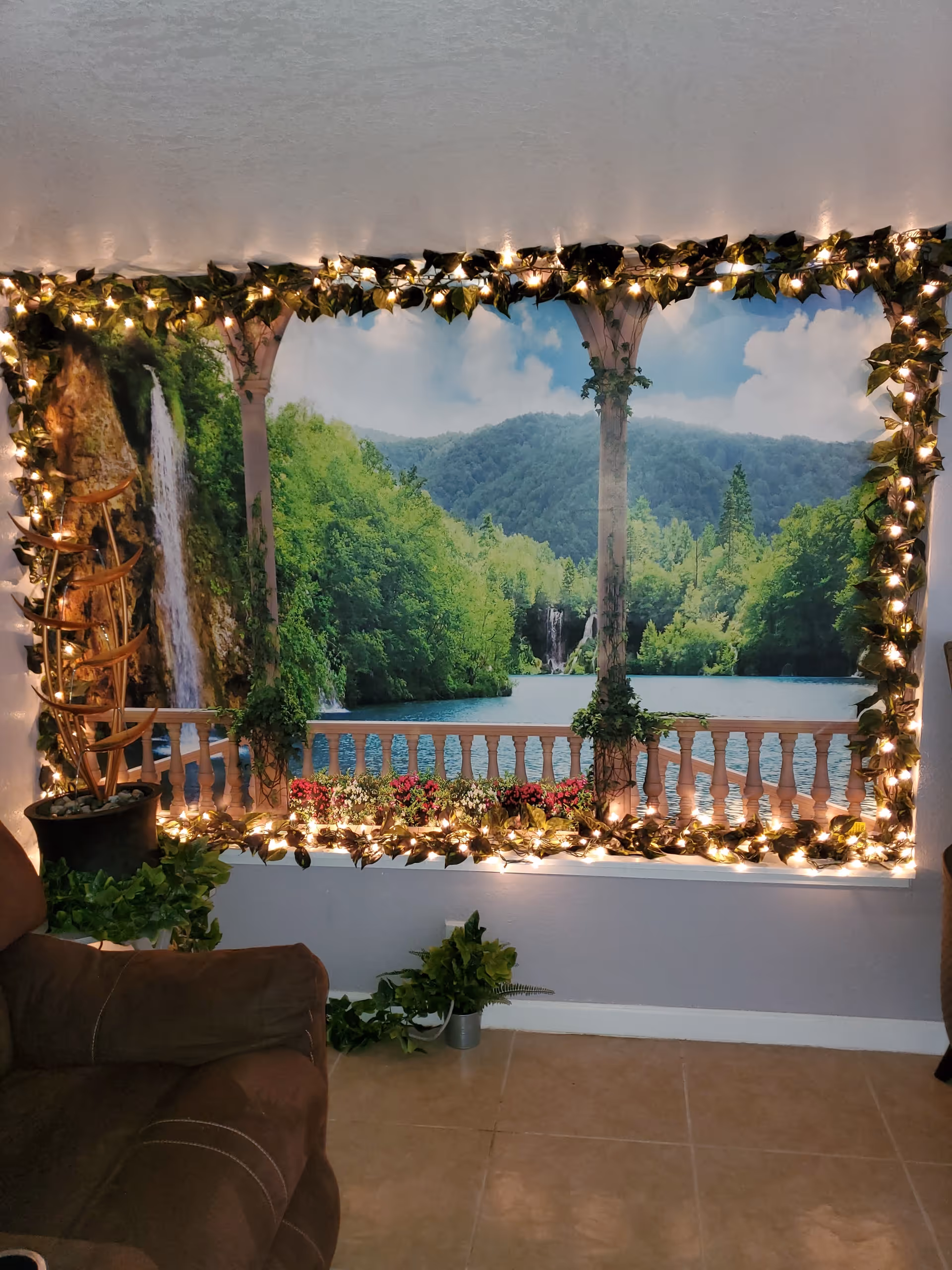 Indoor living area wall decorated with a large scenic mural of a lakeside balcony framed by garland and string lights, with potted plants and a brown recliner in front.