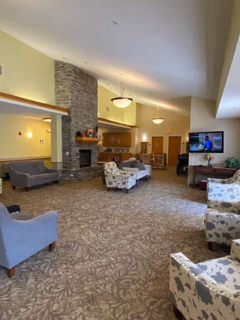 A spacious senior living facility common area with patterned carpet, multiple upholstered chairs and sofas arranged around a stone fireplace. The room has high ceilings with hanging light fixtures, a wall-mounted television, and a small table with flowers and a plant. The walls are painted light yellow and there is a visible kitchen area in the background.