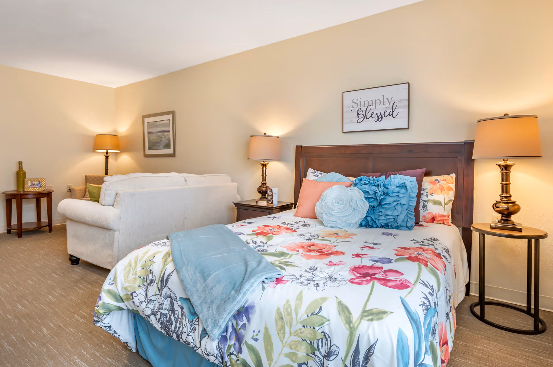 A cozy bedroom with a floral bedspread and decorative pillows on a wooden bed frame. Two bedside tables each hold a lamp with warm lighting. Behind the bed is a wall sign that reads 'Simply Blessed'. In the background, there is a beige couch with a side table holding a bottle and a framed photo, and a floor lamp next to a framed picture on the wall.