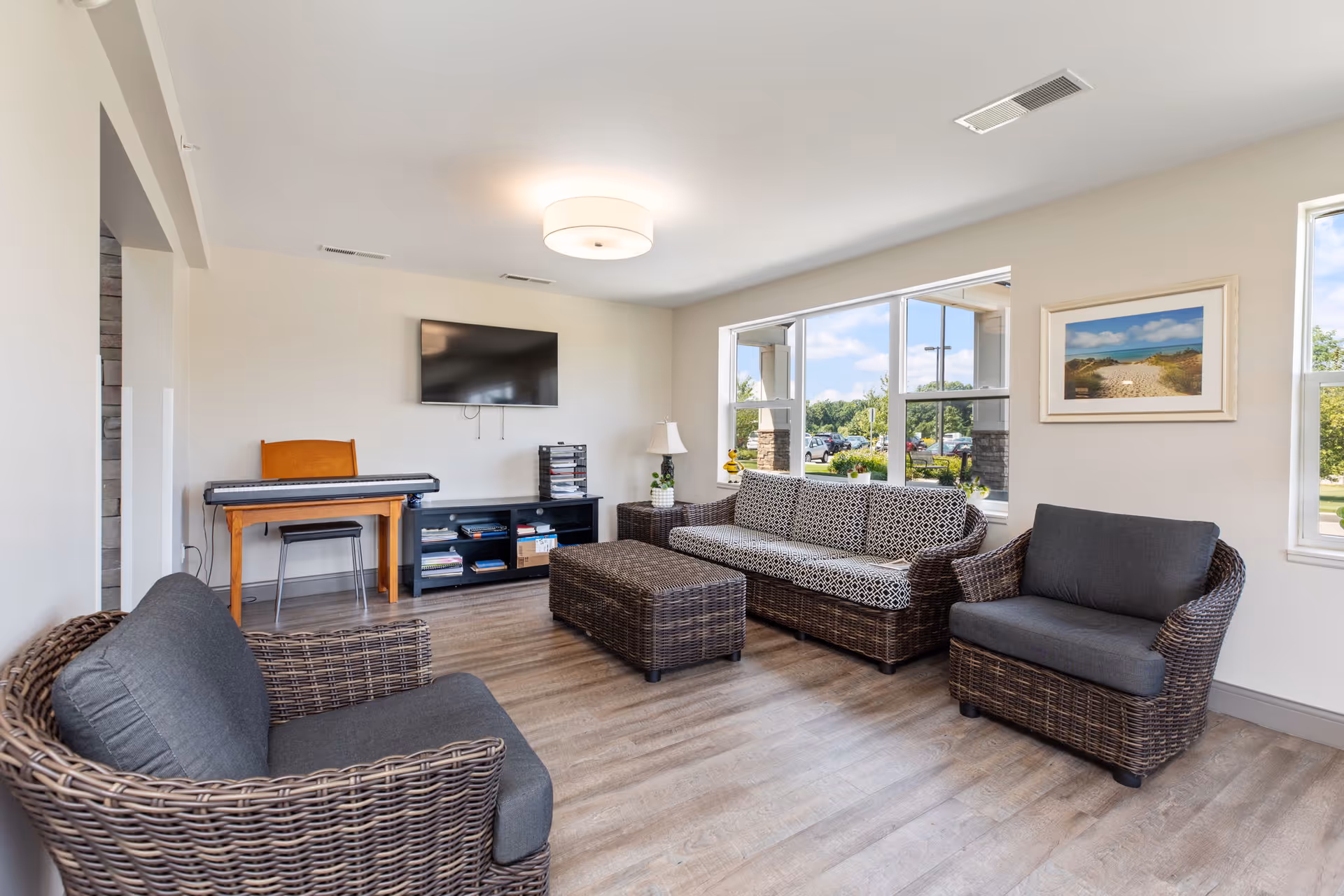 Bright common living room with wicker sofa and armchairs, ottoman, wall-mounted TV, and a keyboard near a large window.