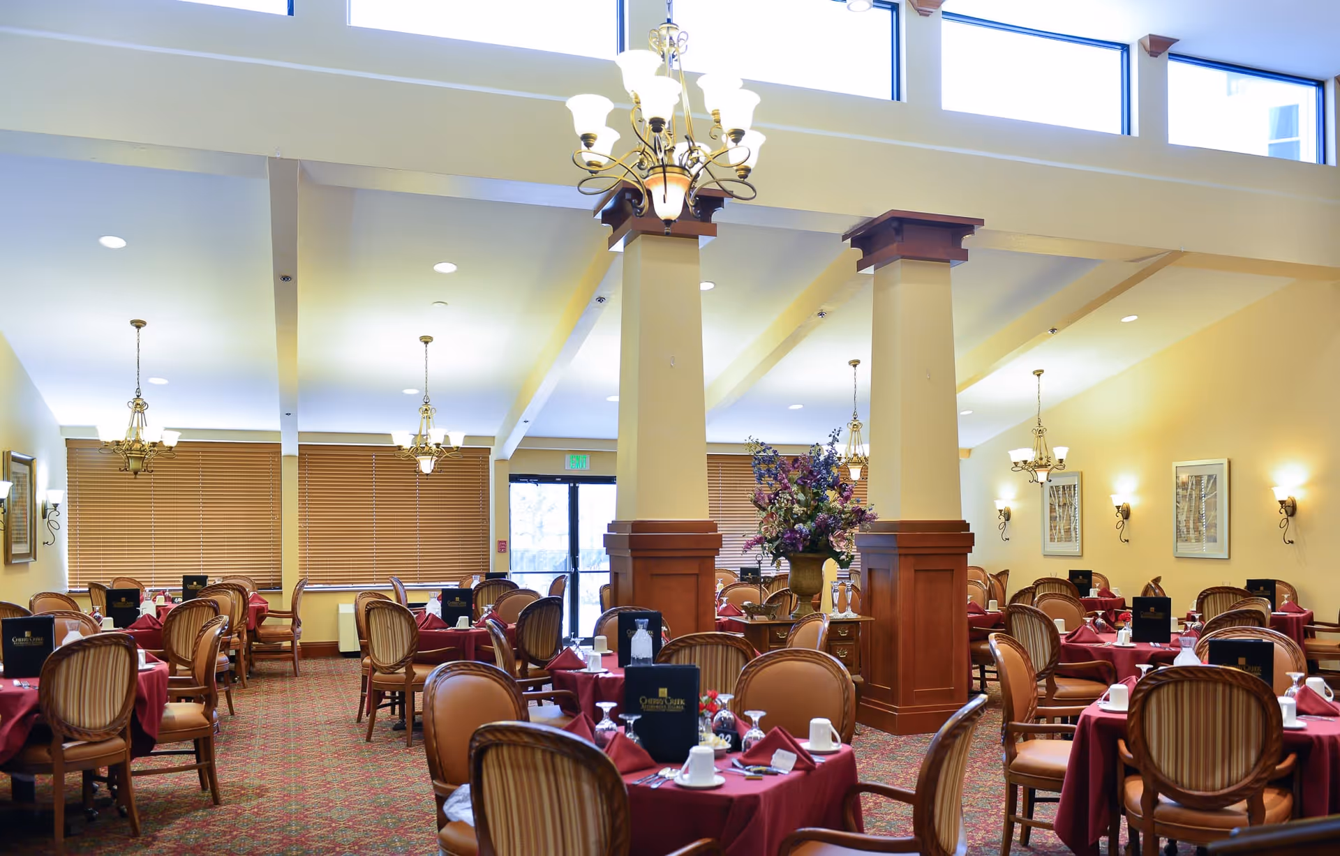 A spacious dining room with multiple round tables covered in red tablecloths, set with white cups, glasses, and silverware. The room features wooden chairs with cushioned seats and backs, large windows with wooden blinds, decorative columns, chandeliers hanging from the ceiling, and a floral arrangement on a small table near the center.