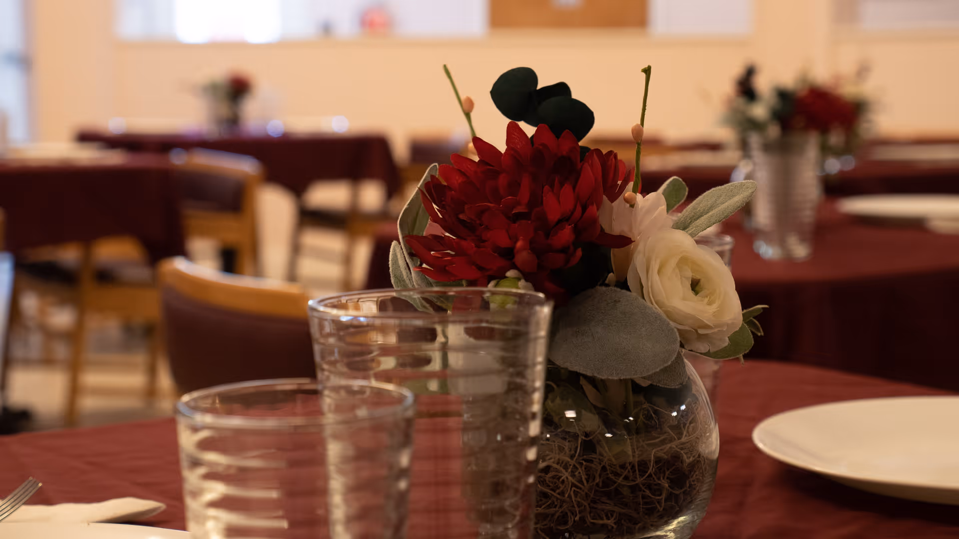 Close-up of a dining table set with a glass vase containing red and white flowers, two empty drinking glasses, and a white plate on a maroon tablecloth. Other tables with similar settings are visible in the softly blurred background.