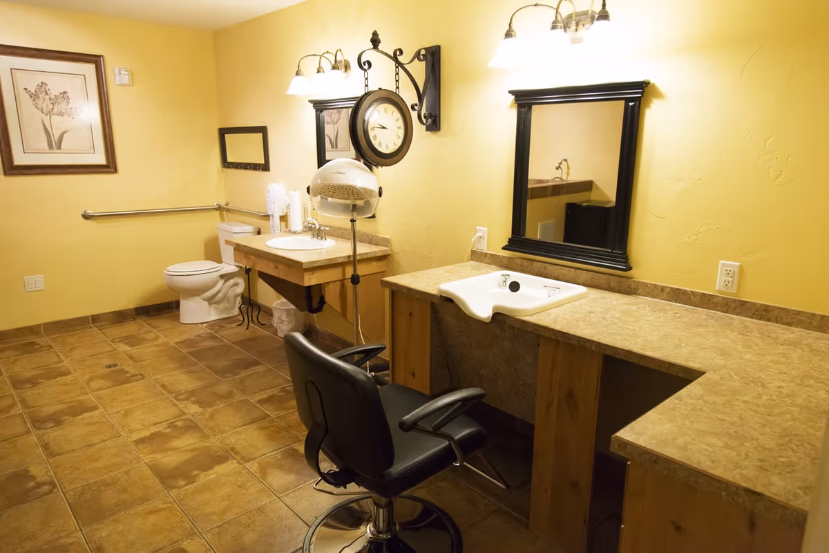 Interior view of a room with yellow walls and tiled floor featuring a toilet, two sinks with mirrors above them, a black salon chair, a wall-mounted clock, and a hair dryer hood, suggesting a combined bathroom and grooming area.