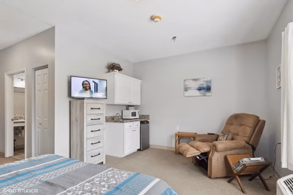 Studio-style room with a bed, small kitchenette and TV on the left and a recliner with side tables on the right.