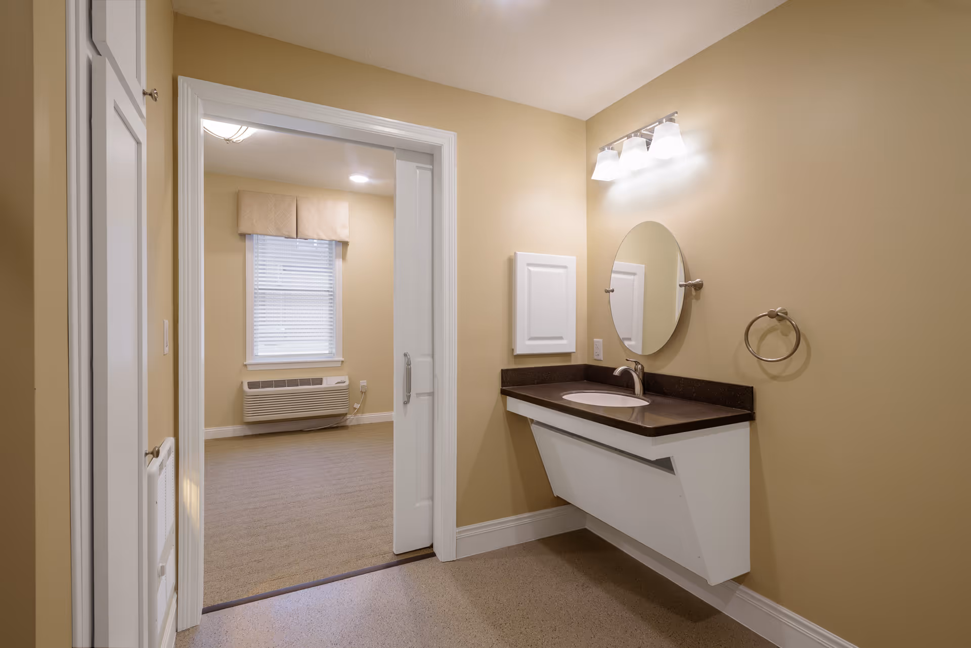 Interior view of a room in Episcopal Church Home showing a beige wall with a mounted sink and countertop, a round mirror above the sink, and a light fixture with three bulbs. There is a towel ring on the wall to the right of the sink. Through an open doorway, a carpeted room with a window covered by a beige valance and an air conditioning unit below the window is visible.