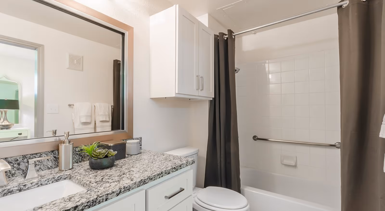 A clean and modern bathroom featuring a granite countertop with a sink, a large mirror above the sink, a white cabinet mounted on the wall, a toilet, and a bathtub with white tiled walls and a dark shower curtain. Towels are visible hanging on a rack reflected in the mirror.