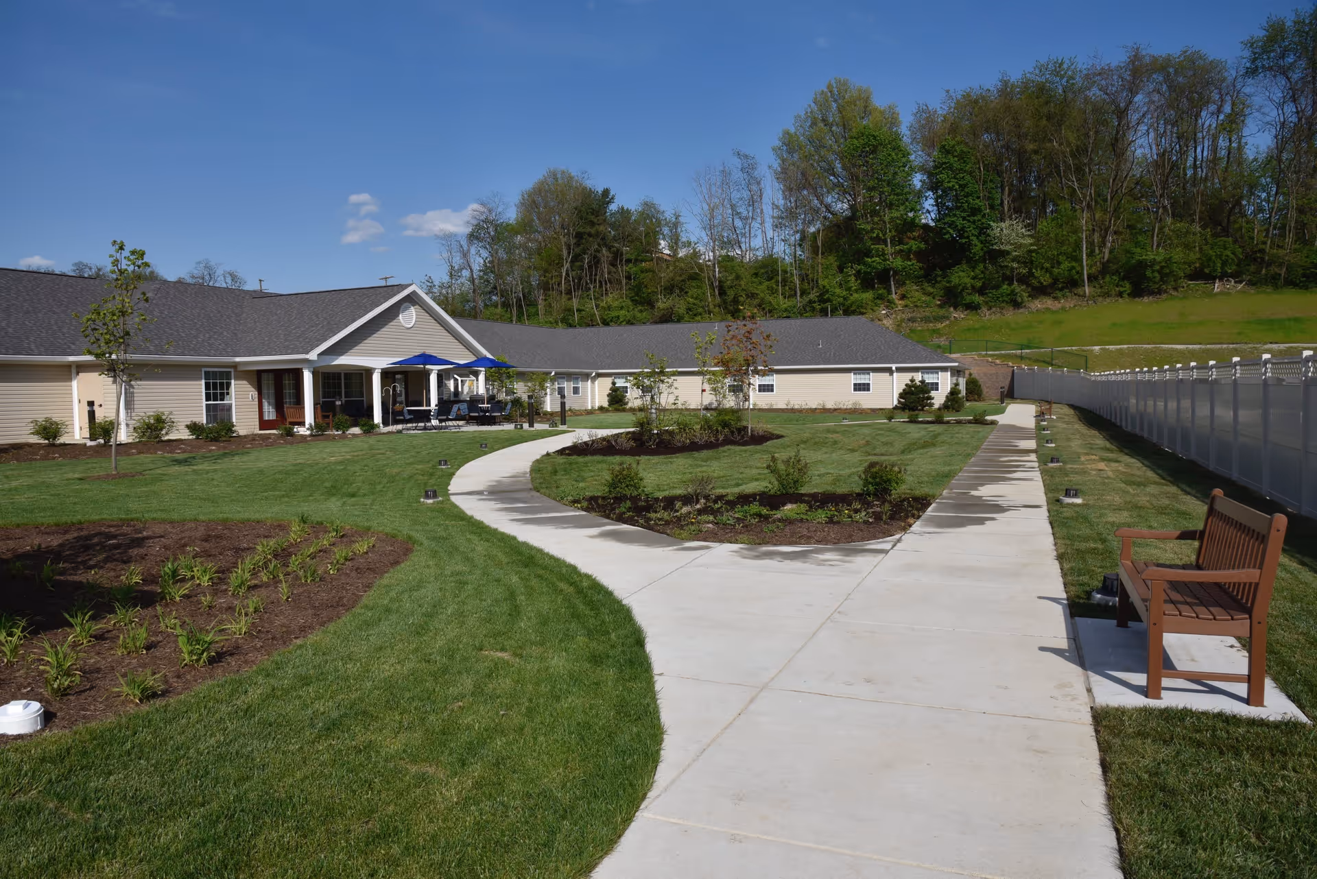 Outdoor view of a senior living facility with a paved walkway curving through well-maintained green lawns and landscaped garden beds. There is a wooden bench on the right side of the path and a building with a covered patio and blue umbrellas in the background. Trees and a clear blue sky are visible beyond the building.