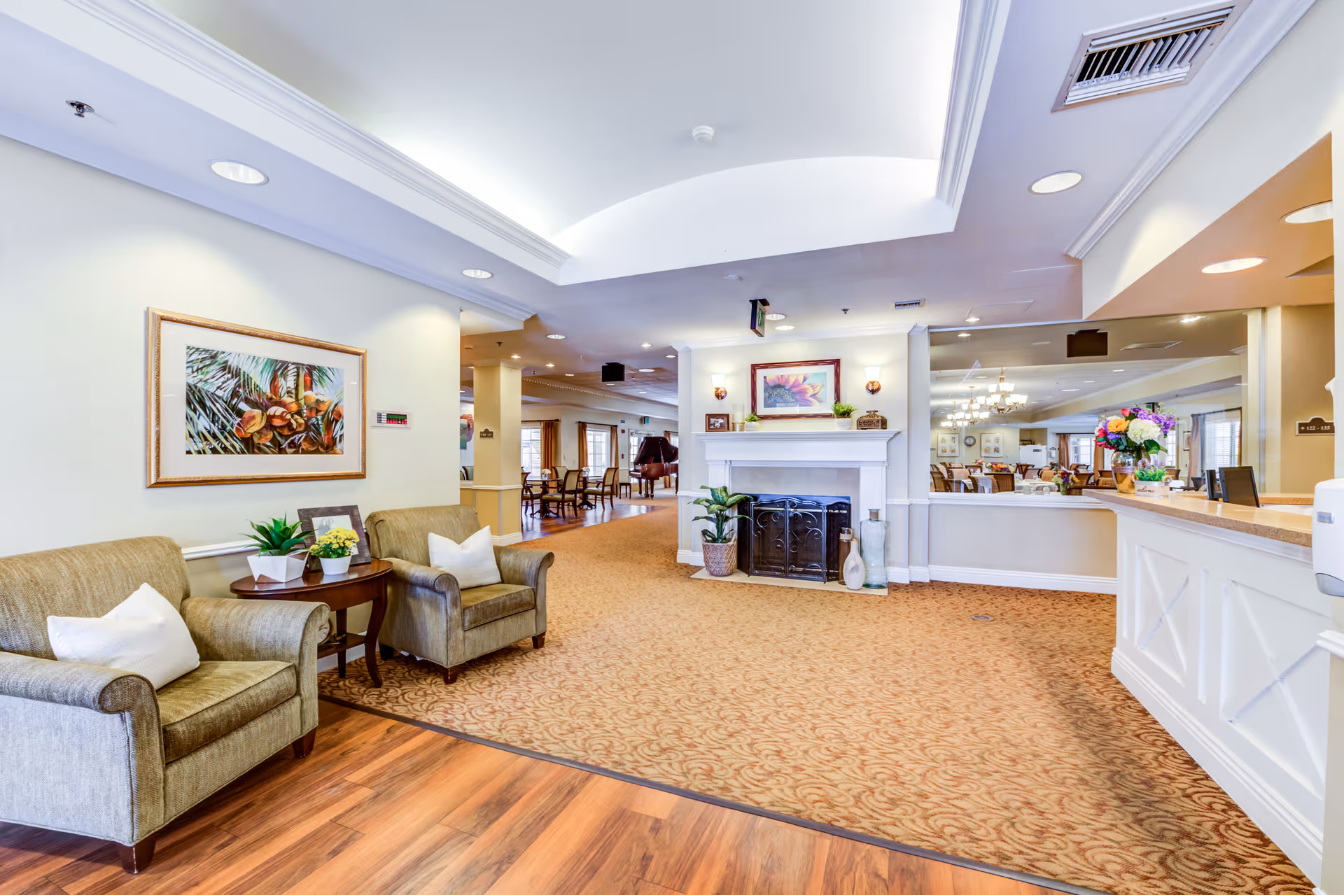 A bright and spacious senior living facility common area with two beige armchairs and a small round table with plants on the left, a white fireplace with decorative items in the center, and a reception desk with flowers on the right. The room has a mix of wood and carpet flooring, recessed lighting, and framed artwork on the walls.
