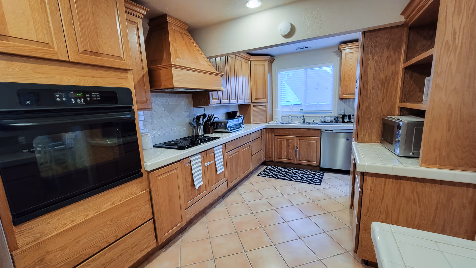 A spacious kitchen with wooden cabinets, a built-in oven, a stovetop with utensils, a dishwasher, a microwave, and a window above the sink letting in natural light. The floor is tiled with a beige pattern and there is a black and white patterned rug in front of the sink.