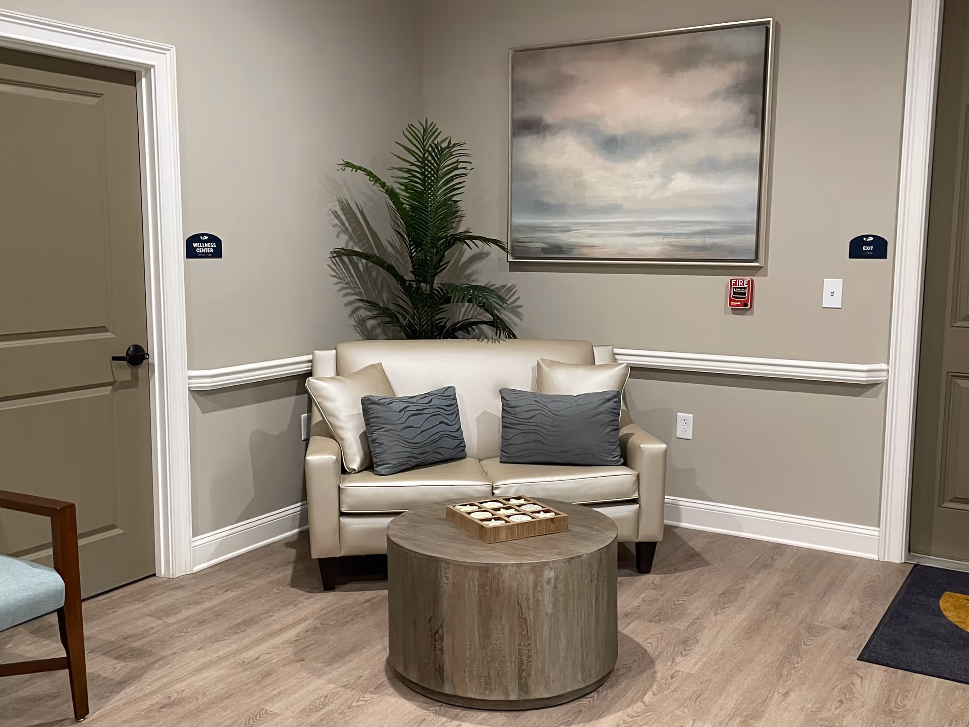 A small seating area in a hallway with a beige loveseat adorned with four pillows, a round wooden coffee table with a decorative item on top, a large potted plant behind the loveseat, and a framed painting of a cloudy sky and ocean on the wall. There are two closed doors on either side labeled 'Wellness Center' and 'Exit'.