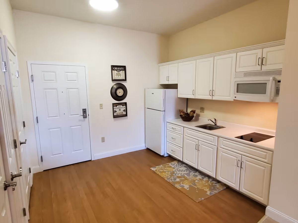 A small kitchen area with white cabinets, a white refrigerator, a microwave, a sink, and an electric stovetop. The floor is wood, and there is a decorative rug in front of the sink. On the wall near the door, there are three framed decorations including a clock. The walls are painted light beige.