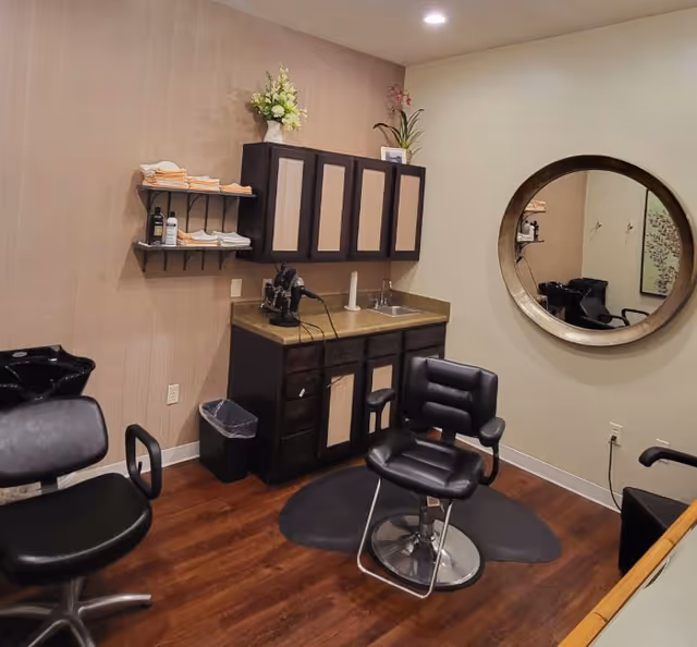 Interior salon room with styling chairs, a round wall mirror, countertop with sink and cabinets, and wood flooring.