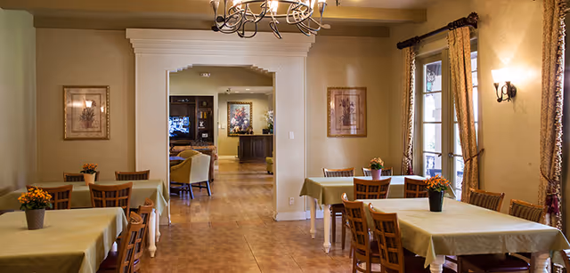 A bright dining room with several tables and wooden chairs covered in green tablecloths, a chandelier overhead, and a view into an adjacent sitting area.