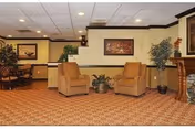 Carpeted communal lounge with two armchairs, potted plants, framed artwork, and a dining table visible in the background.