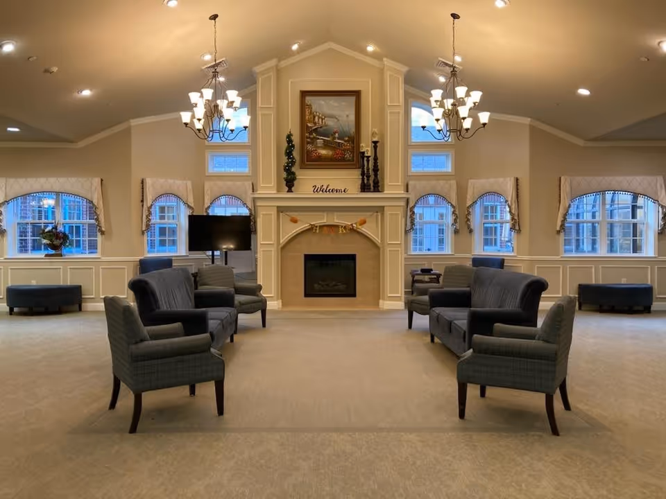 A spacious and elegant living room area with beige walls and carpet, featuring two chandeliers hanging from a vaulted ceiling. The room has multiple windows with decorative valances, allowing natural light to enter. There are several upholstered chairs and sofas arranged facing a central fireplace with a mantel decorated with a painting, plants, and candle holders. A television is positioned near one of the windows.