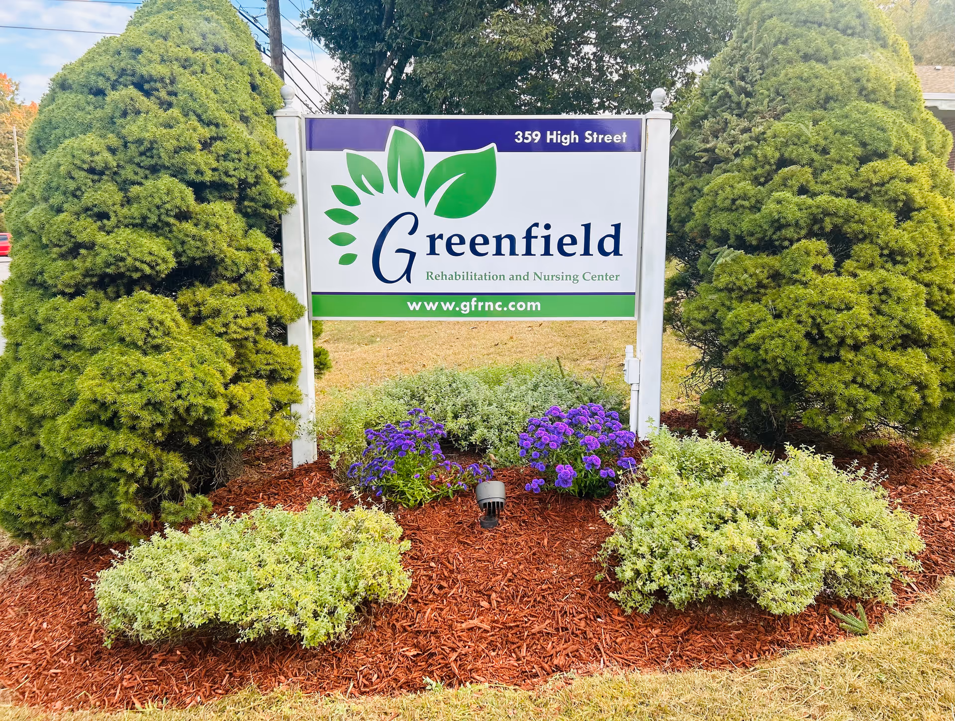 Outdoor sign for Greenfield Rehabilitation and Nursing Center located at 359 High Street, surrounded by green shrubs and purple flowers with a mulched garden bed.
