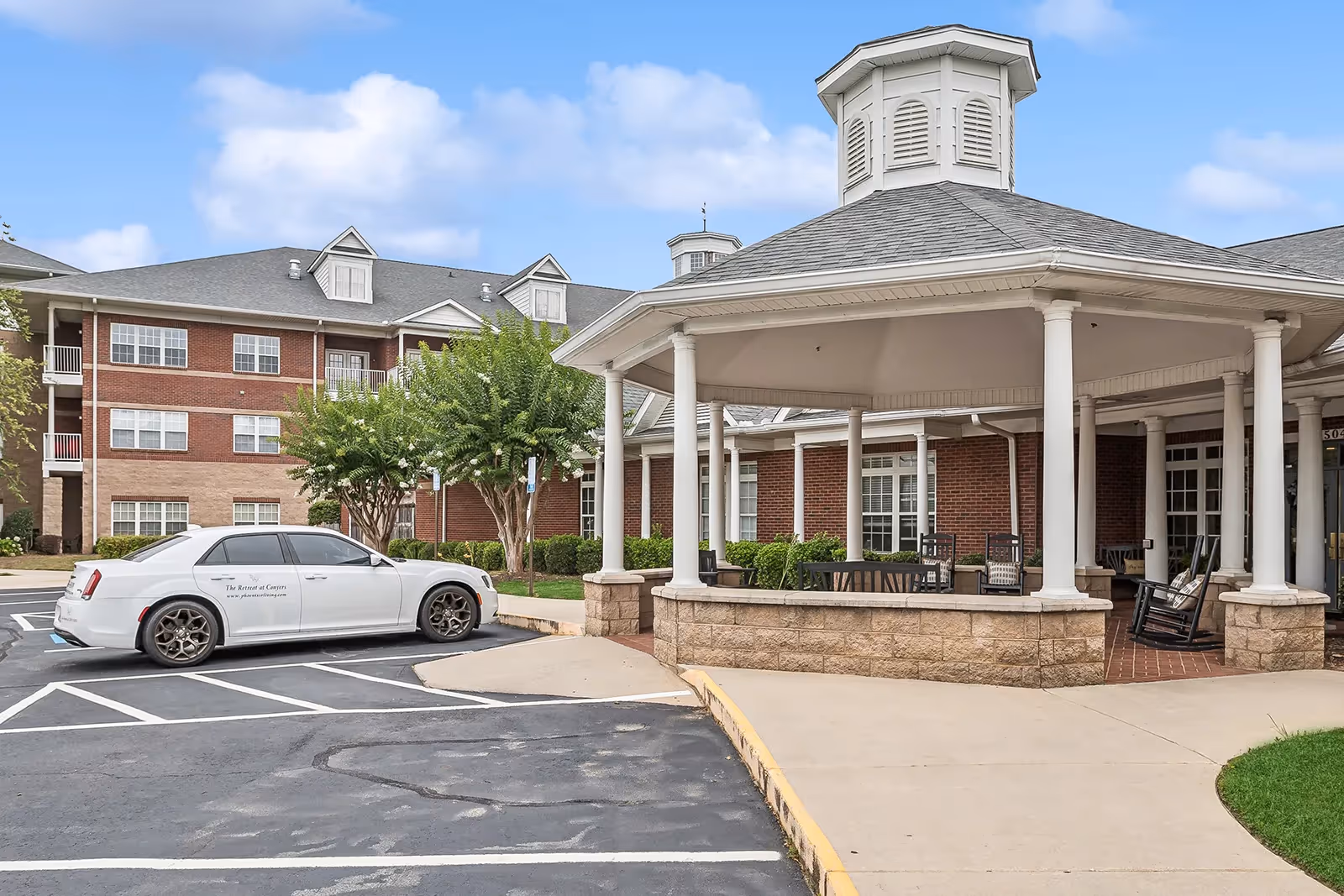 Entrance of a brick senior living building with a covered porte-cochere, parked white car, and outdoor seating.