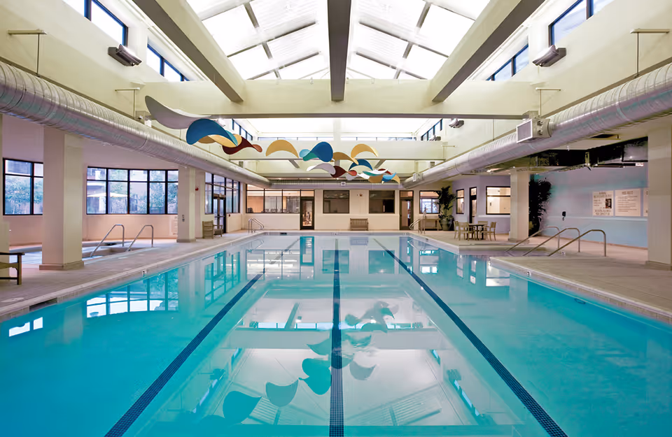 Indoor swimming pool with clear blue water, surrounded by a spacious area with large windows and skylights allowing natural light to fill the space. Colorful abstract decorations hang from the ceiling, and there are handrails and steps for pool access. The area includes seating and tables along the sides.