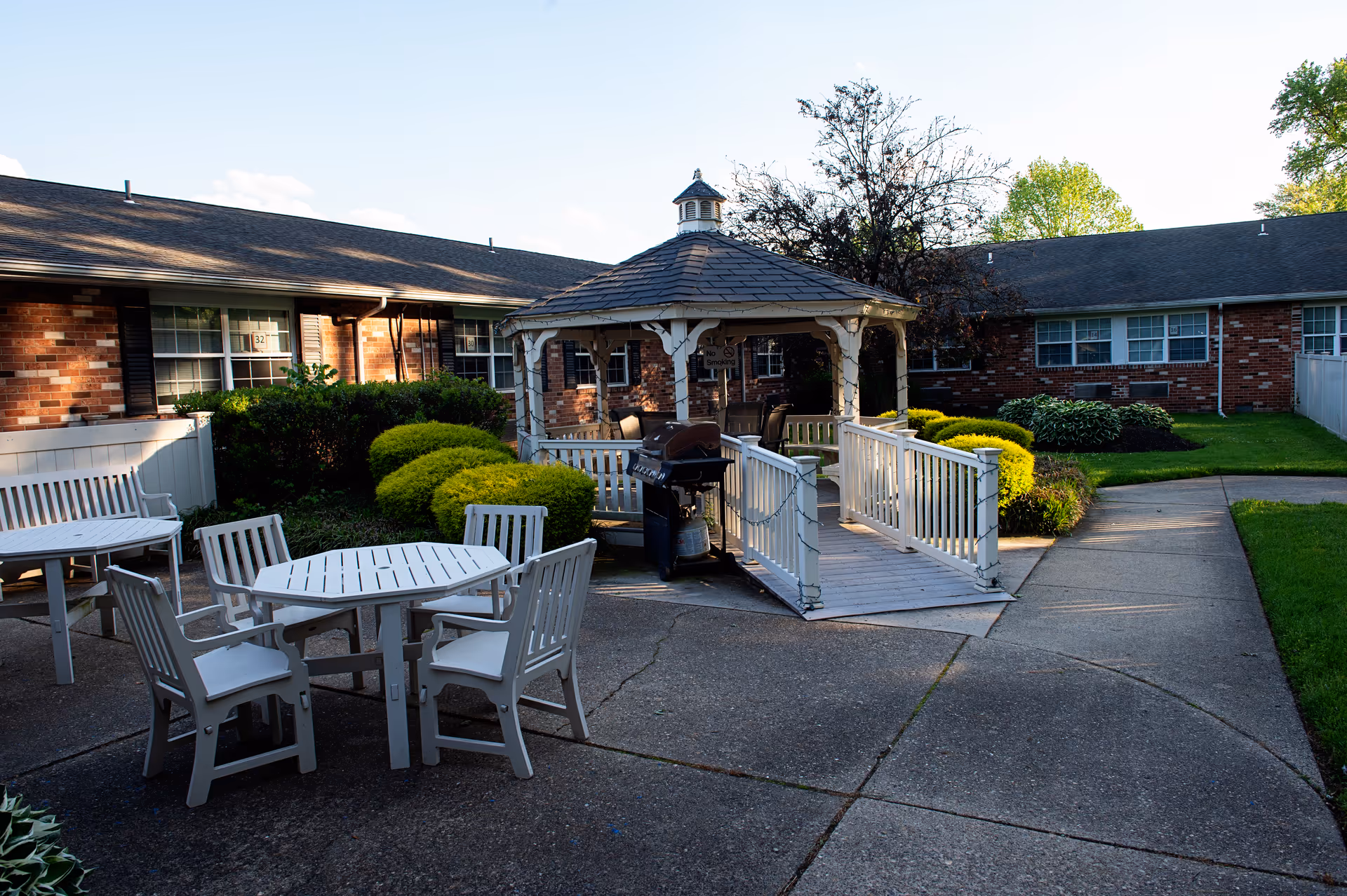 A landscaped courtyard featuring a central gazebo, patio tables and chairs, a grill, and surrounding brick residential buildings.