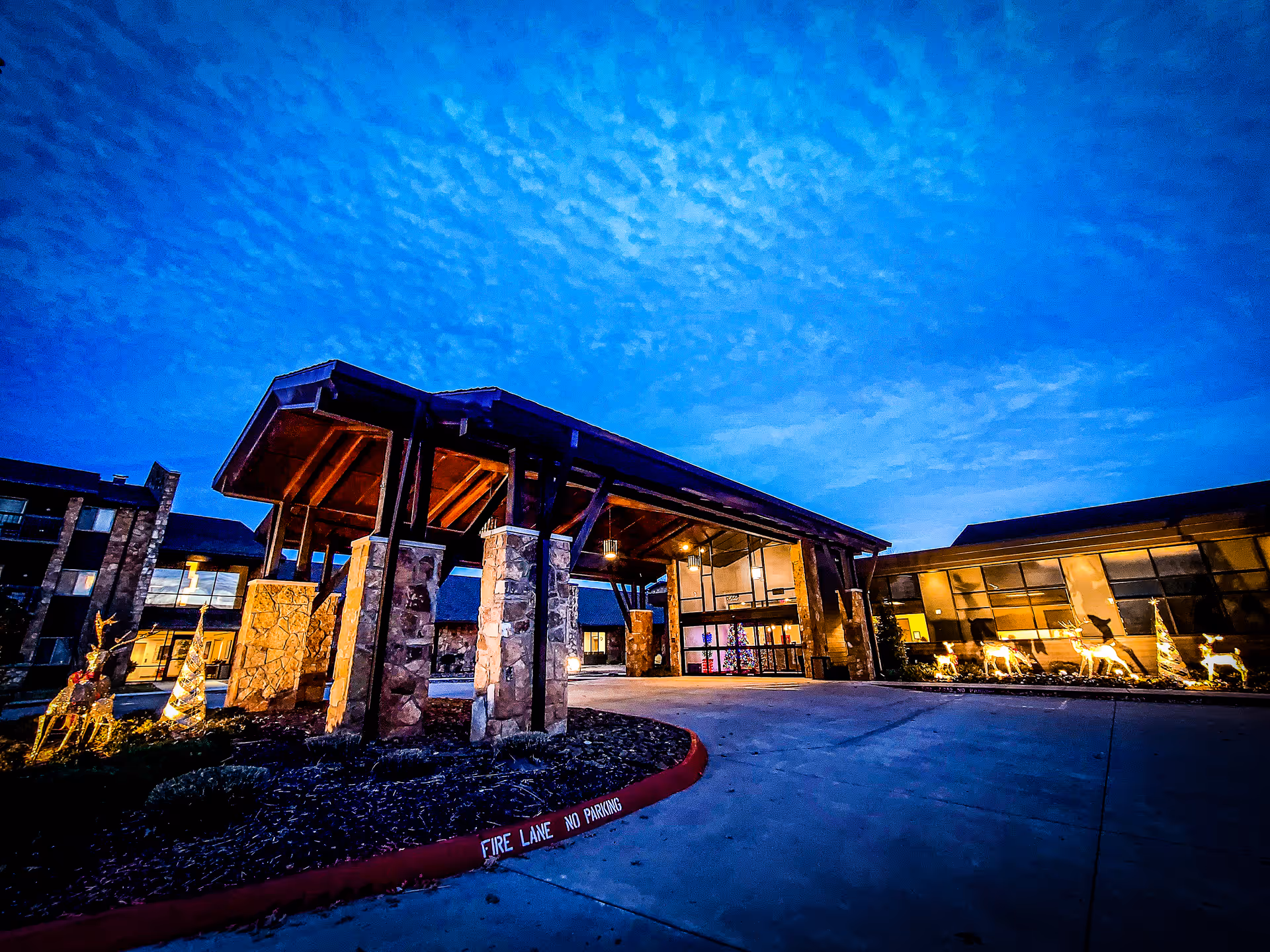 Front entrance of a building with a large wood-and-stone covered porte-cochere lit at dusk and holiday decorations.